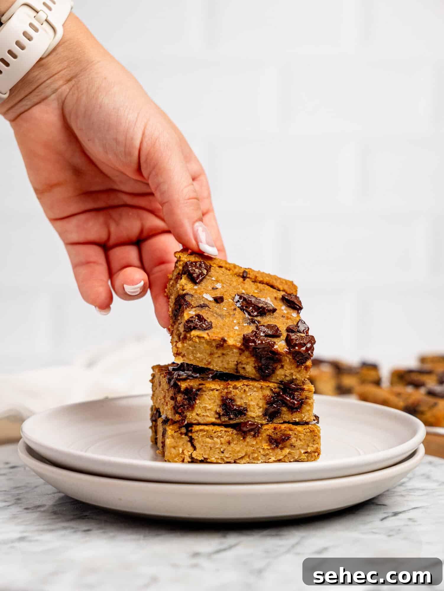 Gooey Chickpea Cookie Bars 8 Three chickpea cookie bars stacked on top of each other on a white plate, a hand is picking up the one on top.