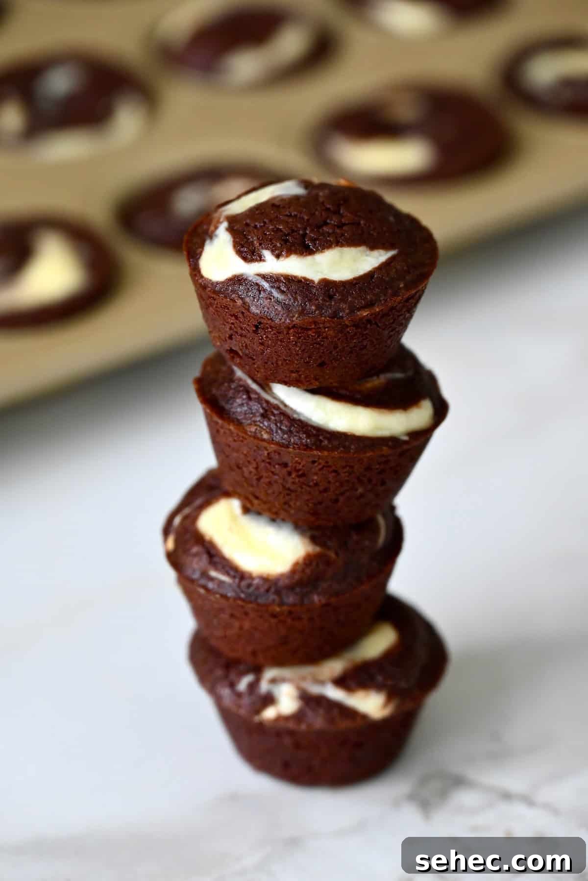 Cheesecake brownie bites are stacked on a marble counter, with a mini muffin pan in the background.
