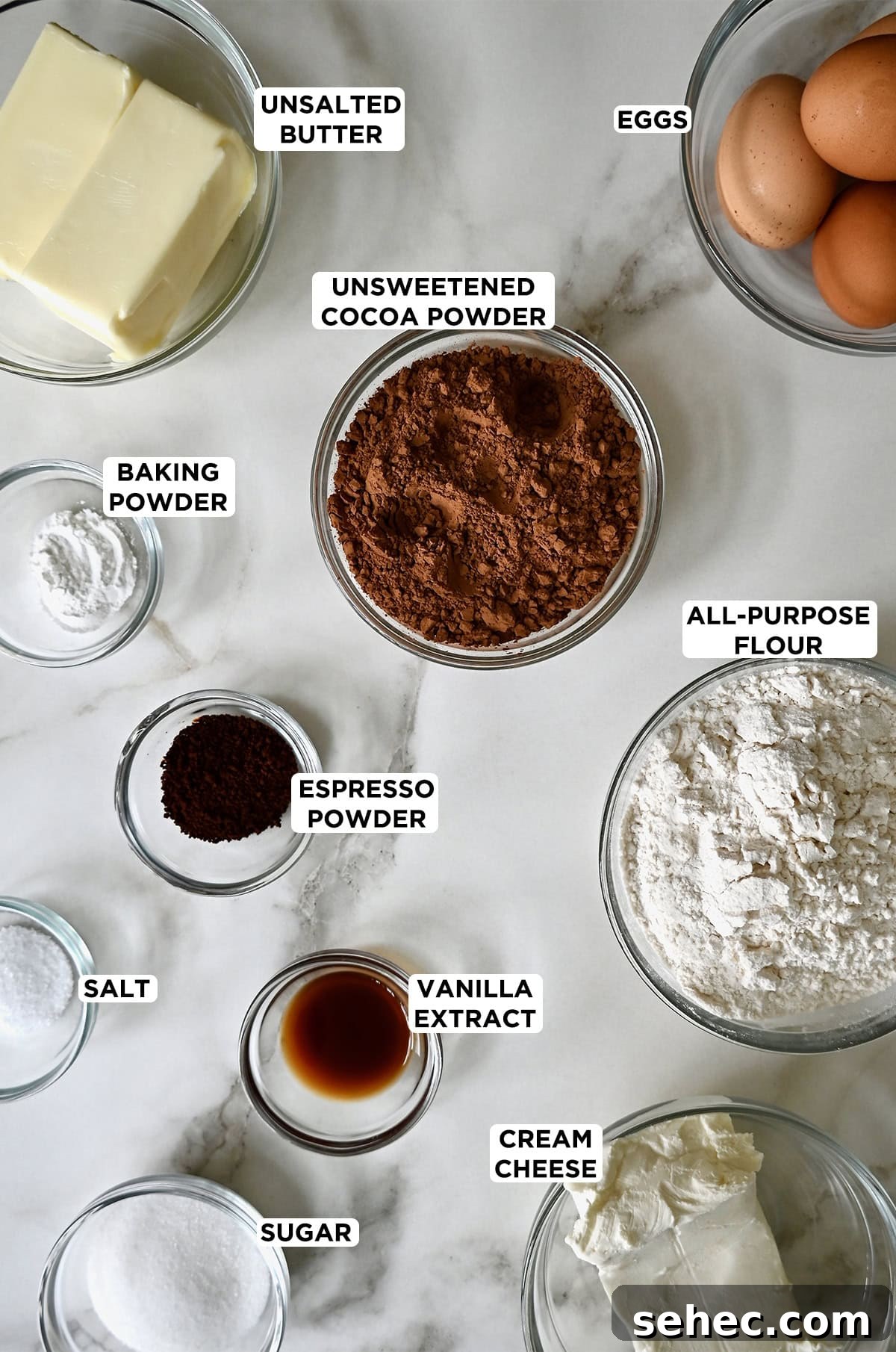Clear glass bowls are filled with the ingredients for cheesecake brownie bites, including sugar, cream cheese, vanilla, flour, salt, espresso powder, baking powder, butter, eggs and cocoa powder.