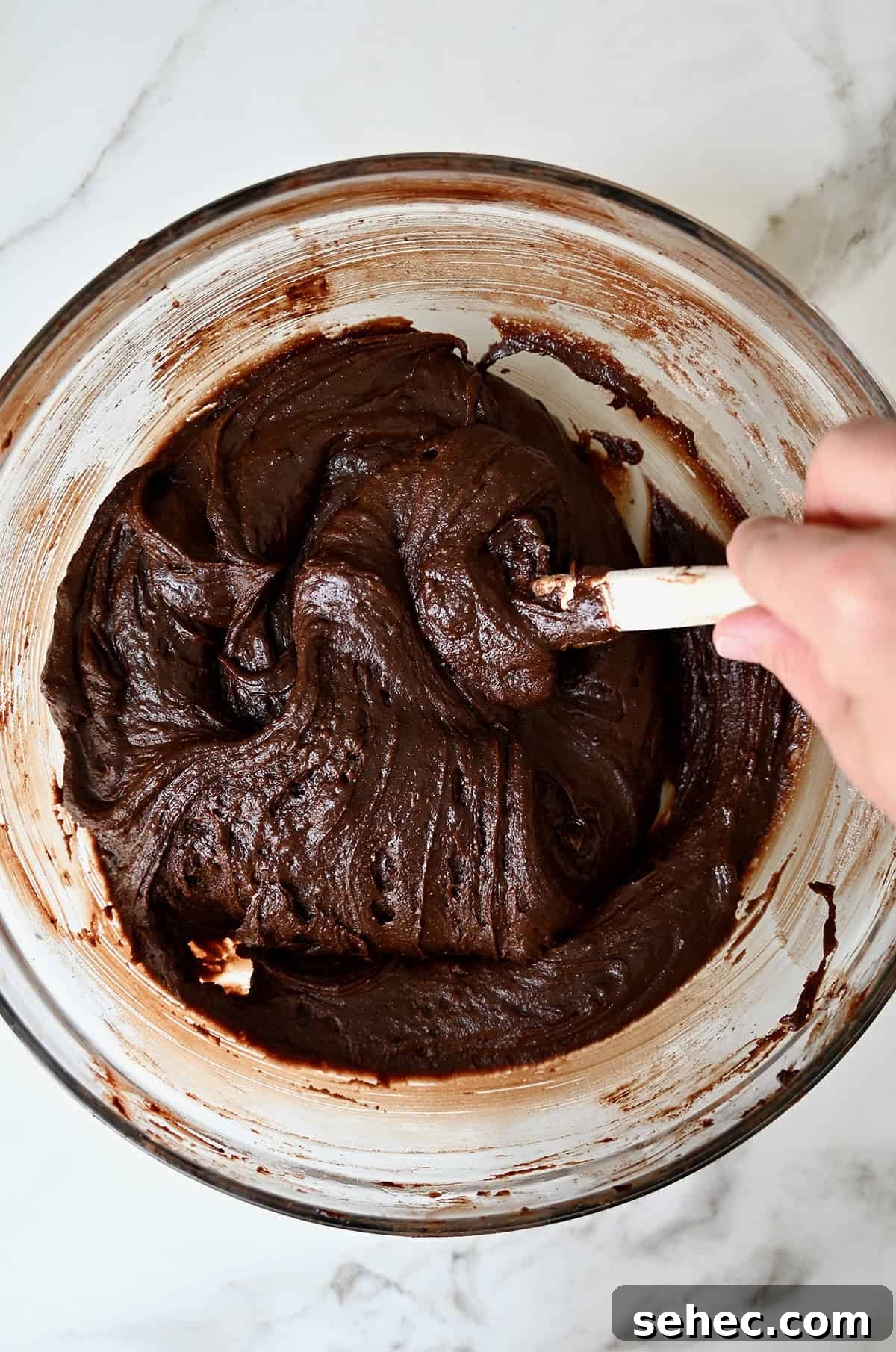 Brownie batter is being mixed with a spatula in a glass bowl.