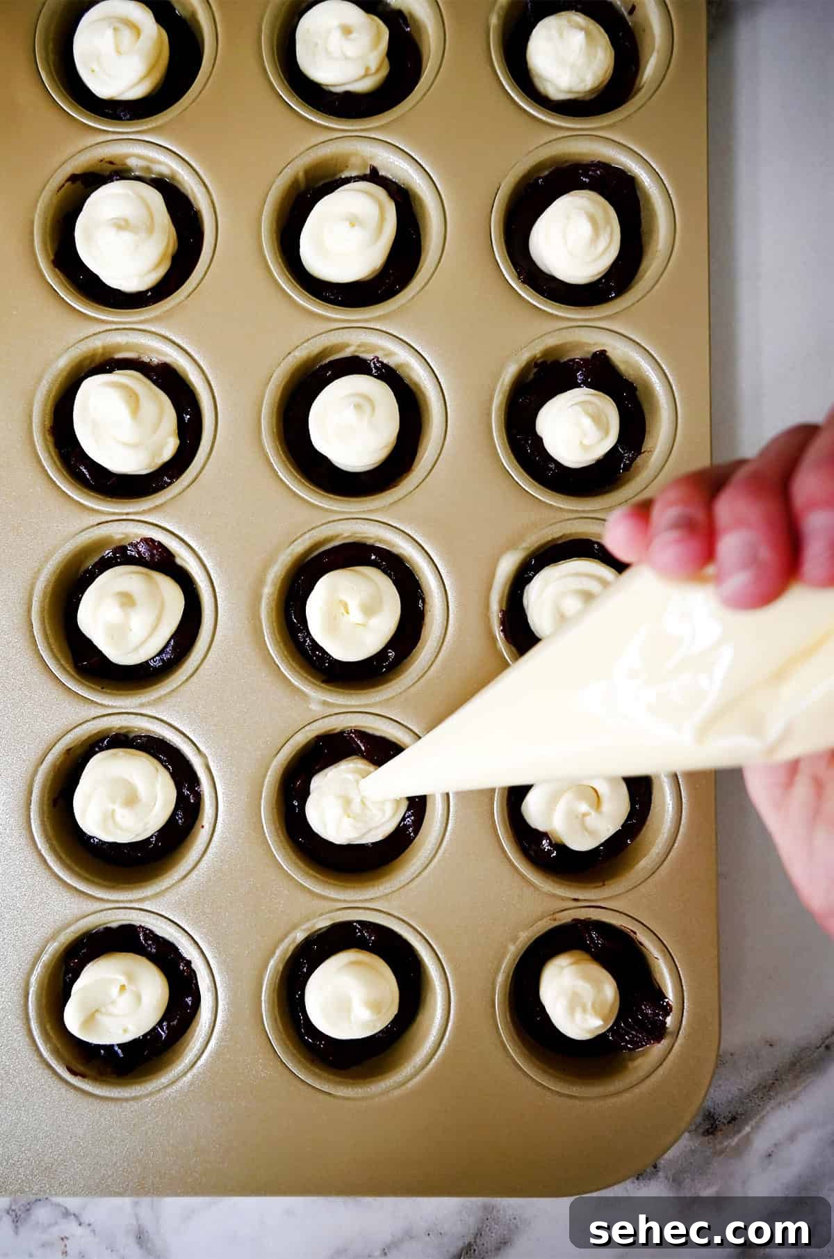 Cheesecake filling is piped onto brownie batter in a mini muffin pan.