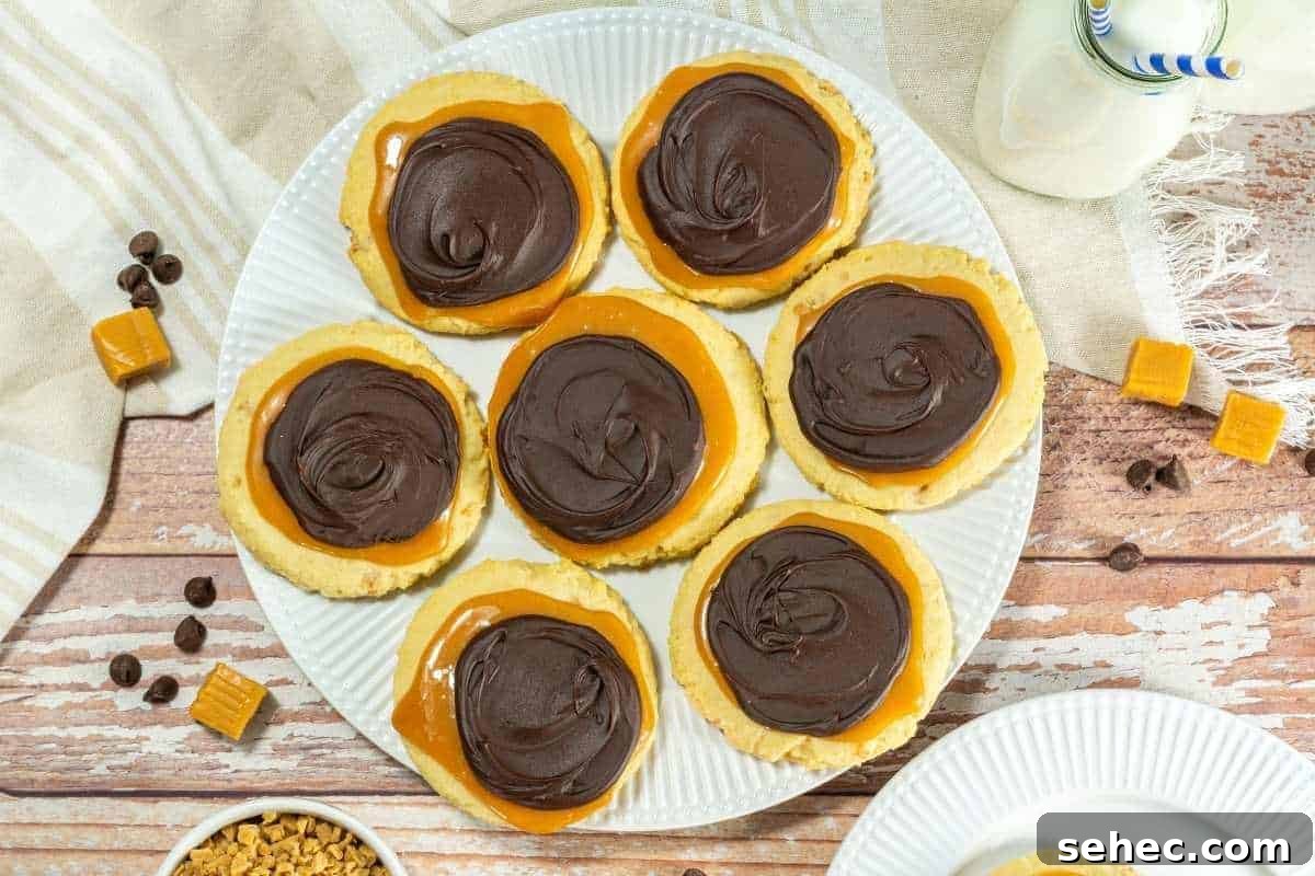 17 Buttery Fall Cookie Recipes for Cozy Shorter Days 5 A plate of Crumbl Copycat Twix cookies surrounded by chocolate chips, caramel pieces, a glass of milk, and a beige cloth on a wooden surface.