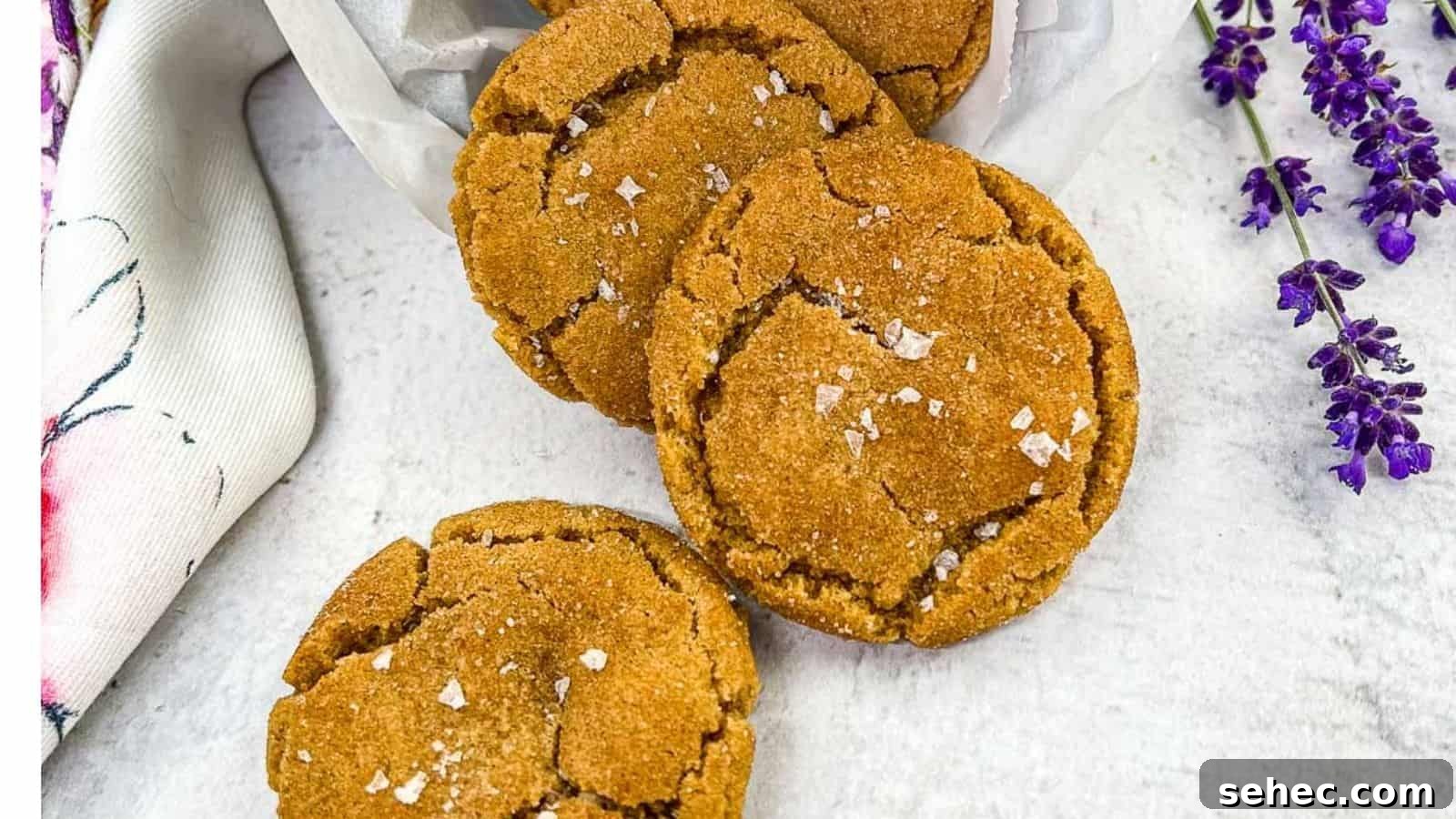 17 Buttery Fall Cookie Recipes for Cozy Shorter Days 9 Three brown butter snickerdoodle cookies arranged on a white surface, sprinkled with sea salt flakes, with a white cloth and purple flowers on the side.