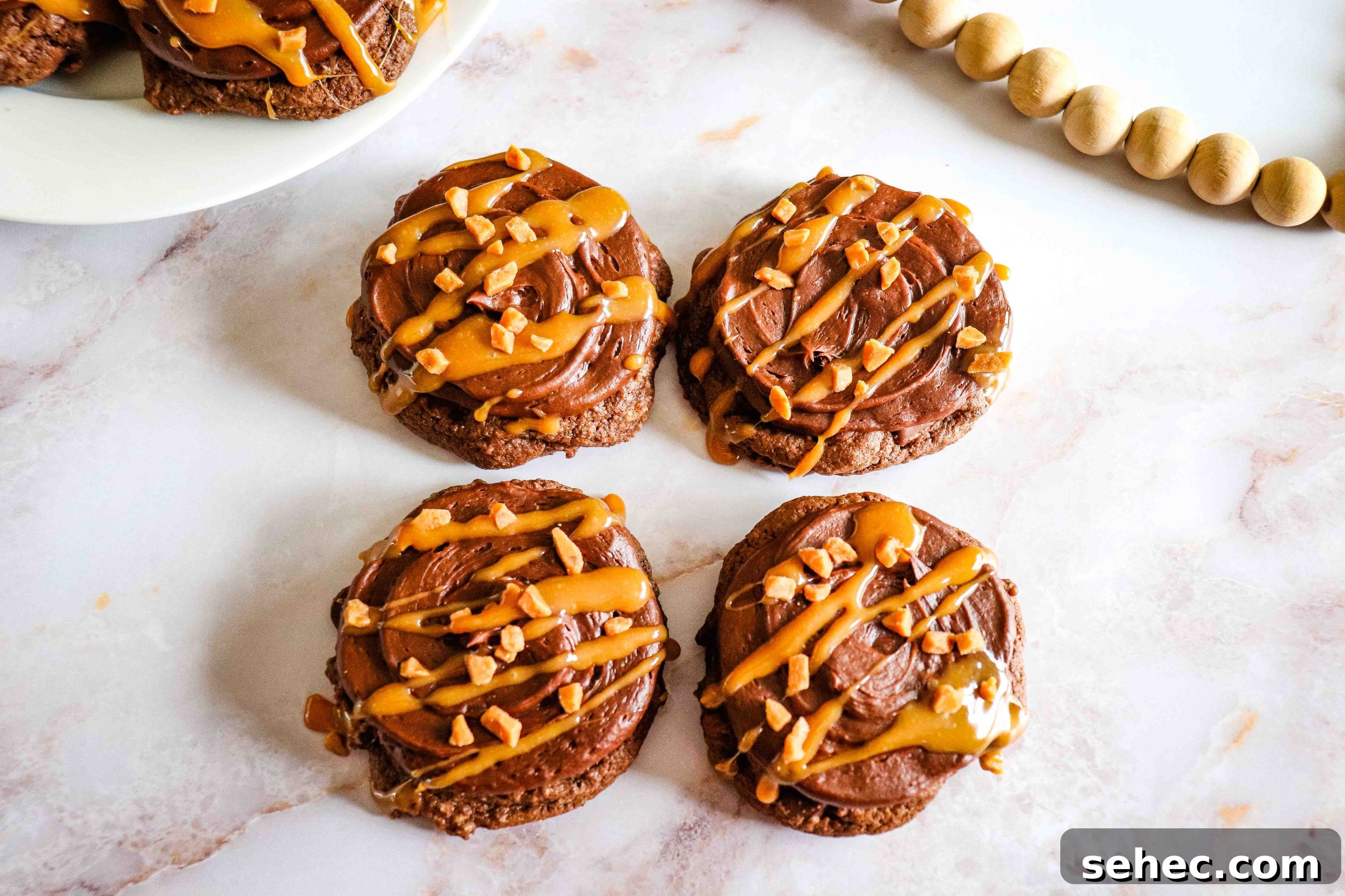 17 Buttery Fall Cookie Recipes for Cozy Shorter Days 10 Chocolate Caramel Cookies with frosting, caramel drizzle, and chopped nuts on a white marble surface.