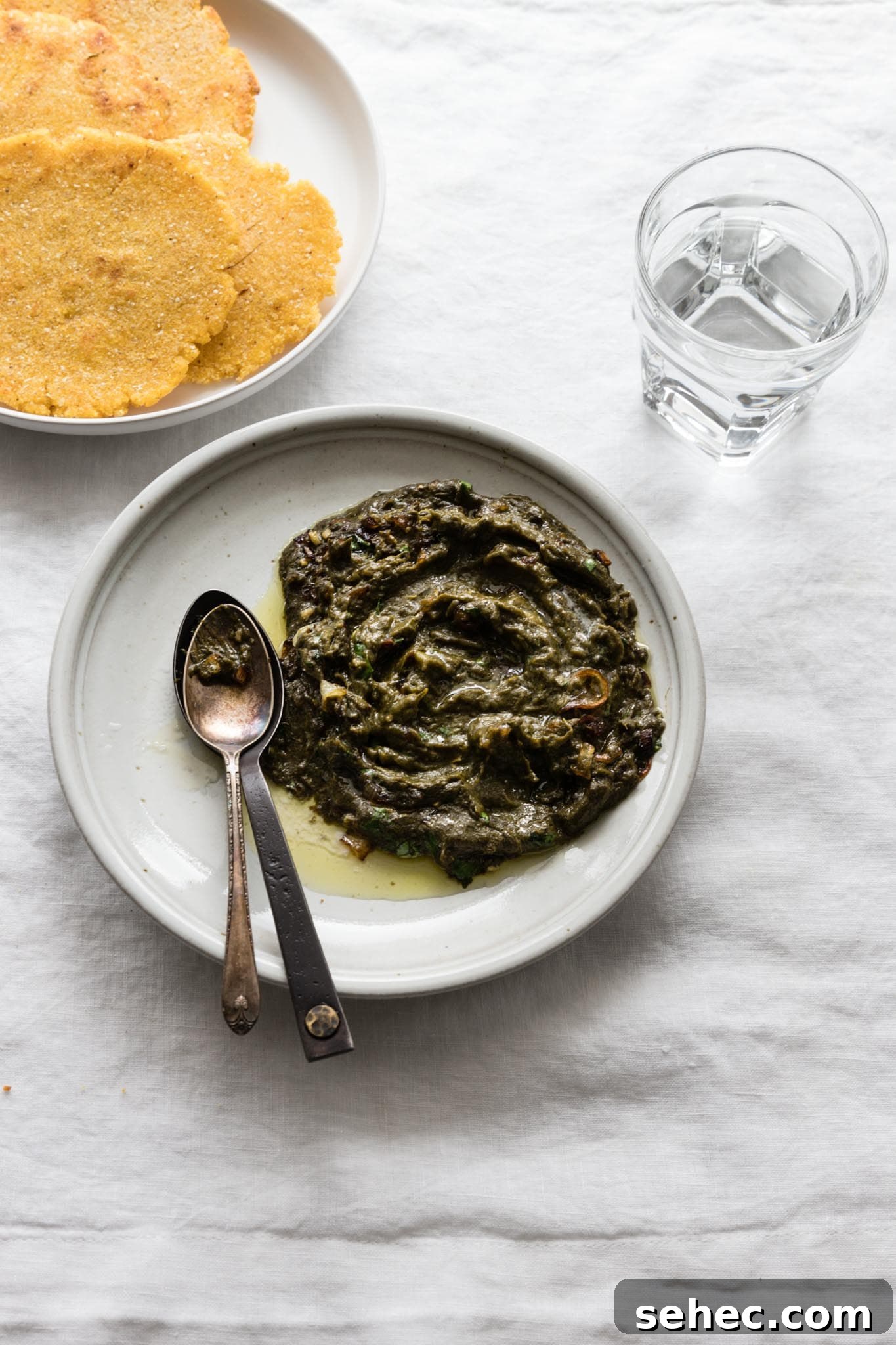 Sarson Ka Saag Recipe Stovetop and Instant Pot 14 Easy Sarson Ka Saag on a plate with a couple spoons on the side served with makai ki roti (cornmeal flatbread) and a glass of water