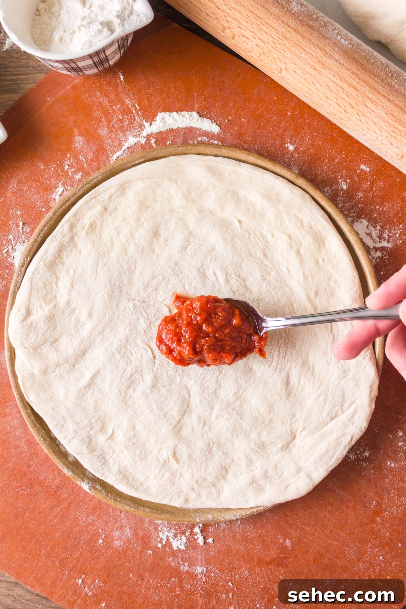 Crispy Chewy Sourdough Pizza Crust Recipe 14 Pizza sauce being spread on a rolled homemade sourdough pizza crust