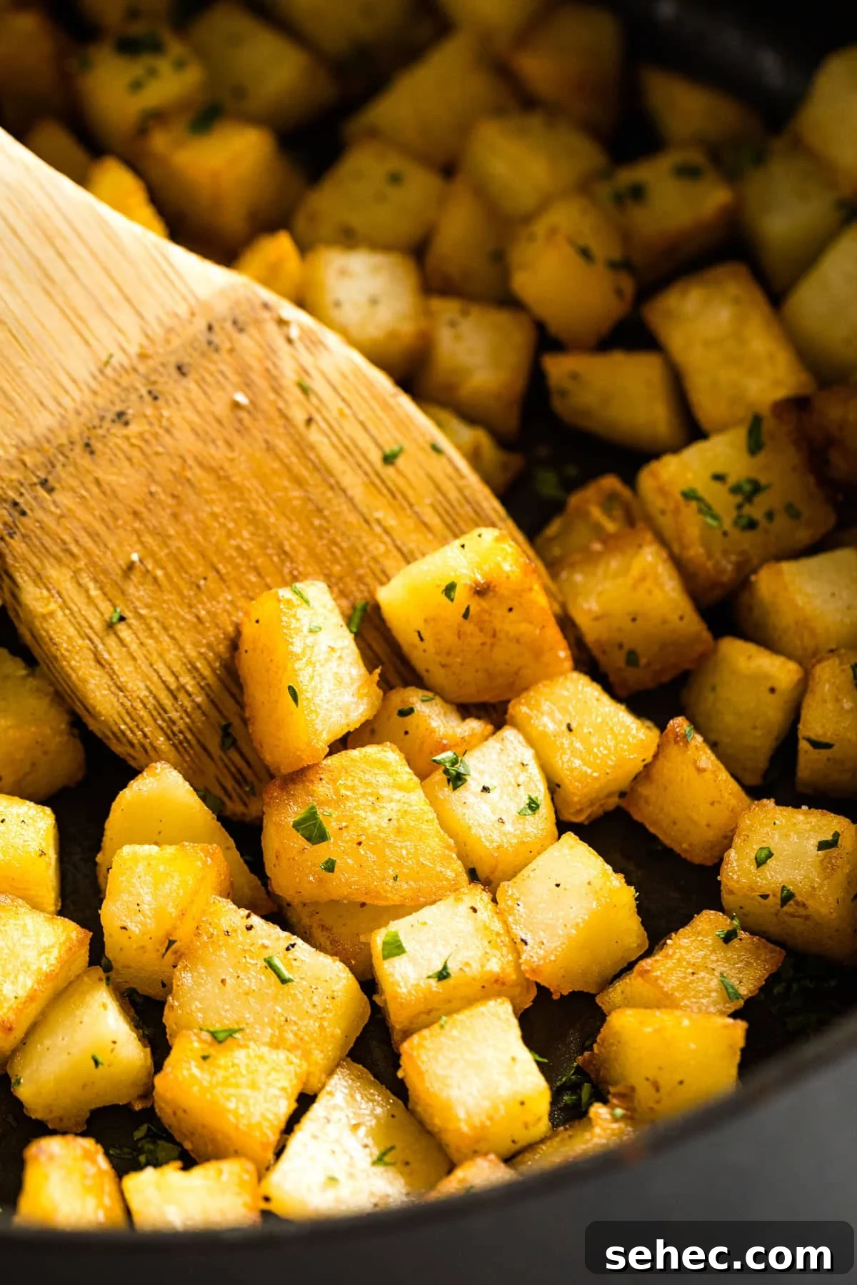 Crispy Fried Potatoes Recipe 2 A close up of fried potatoes in a pan with a wooden spoon in it.