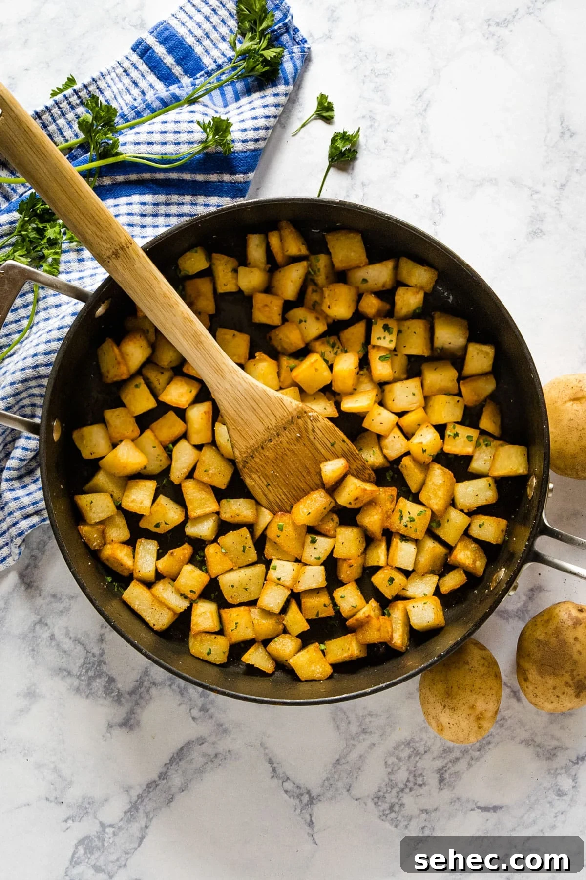 Crispy Fried Potatoes Recipe 3 An overhead image of a pan of fried potatoes with a wooden spoon in it and potatoes and a towel on the side.