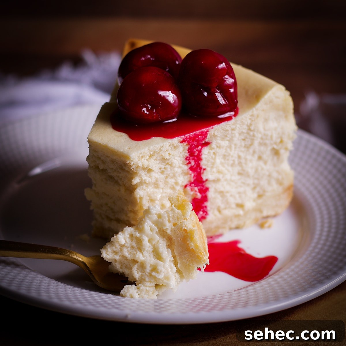 A close up shot of a bite of creamy New York Cheesecake on a fork with the rest of the slice of cheesecake behind it.
