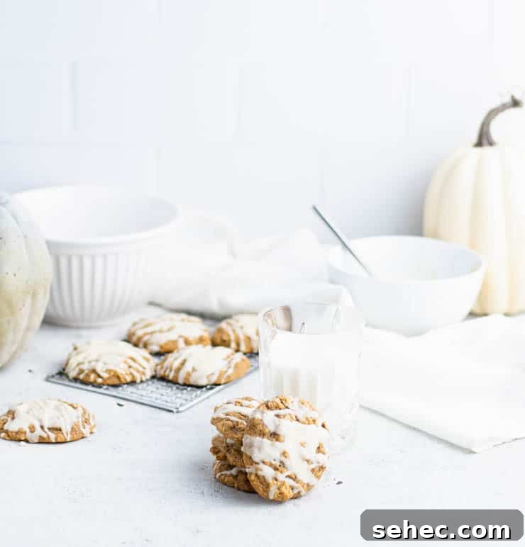 Pumpkin Oatmeal Cookies with Brown Butter Icing 2 a stack of iced pumpkin cookies next to a short glass of milk with additional cookies, bowls and white and green pumpkins in the background against a tiled backdrop.