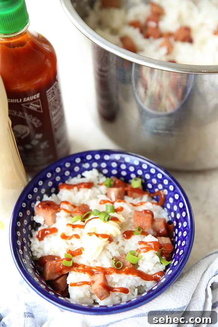 A pot of spam and rice, a bottle of sriracha sauce, and a dark blue bowl with white polka dots filled with rice and spam, drizzled with sriracha sauce, and topped with mayonnaise and green onions. A striped tea towel is wrapped around the bottom of the bowl.