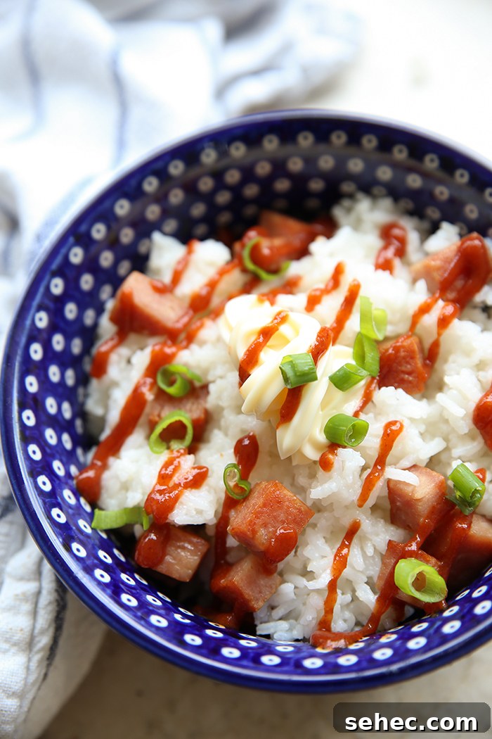Spam and rice topped with mayonnaise, sriracha sauce, chopped green onions in a peacock patterned Polish pottery bowl. A striped tea towel is wrapped around the left of the bowl.