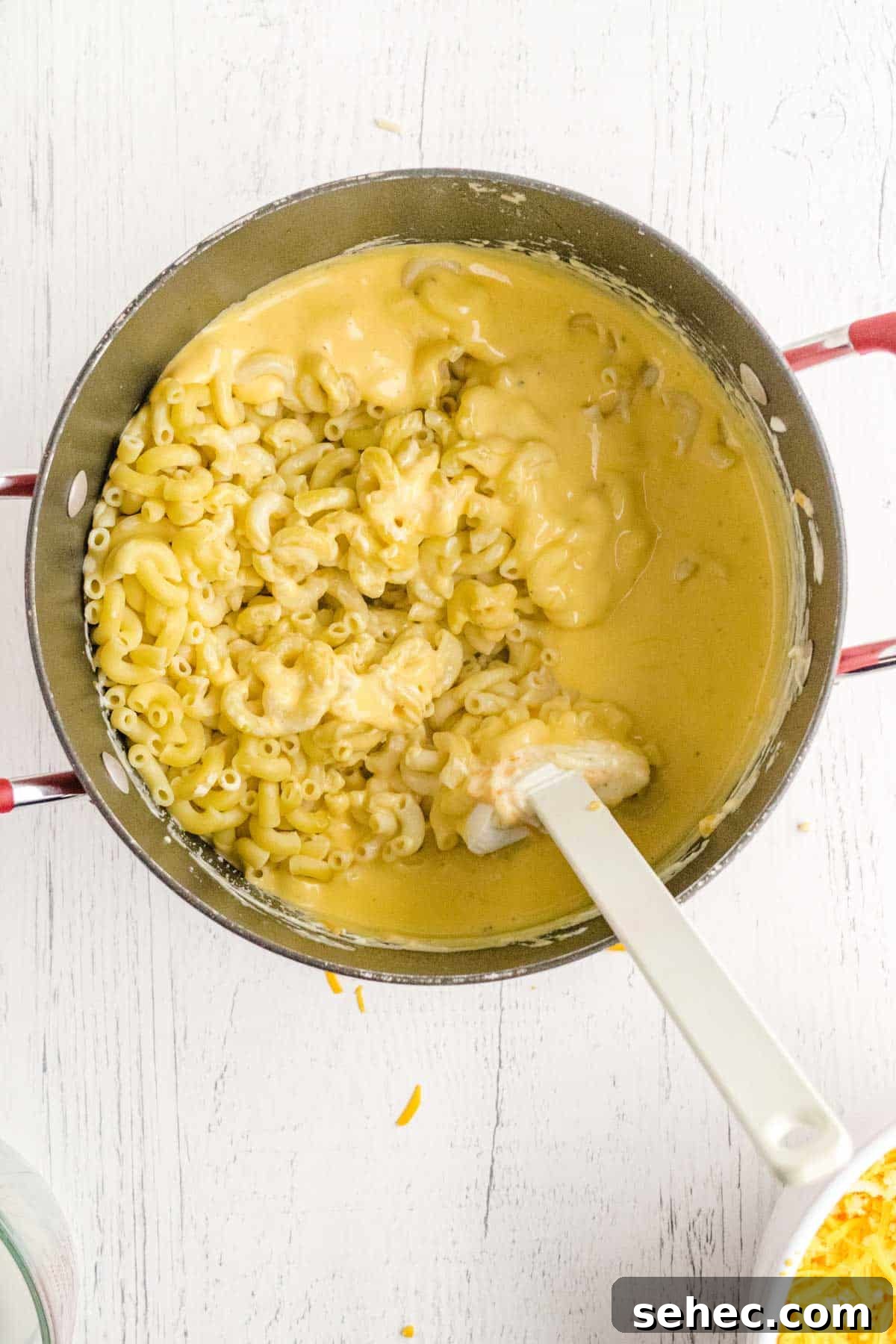 Creamy Eggless Mac and Cheese Recipe 11 A pot of cooked elbow macaroni being mixed with creamy cheese sauce using a white spatula on a white wooden surface.