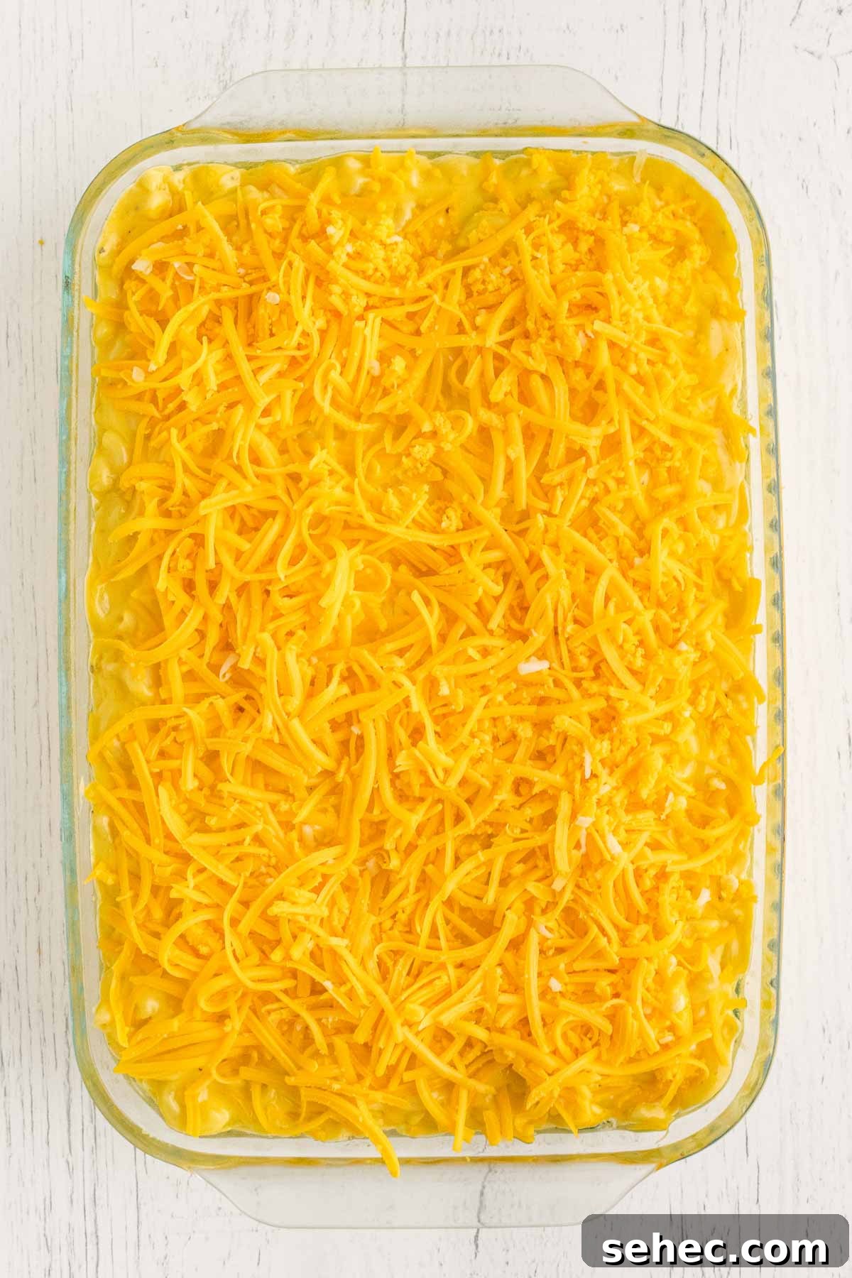 Creamy Eggless Mac and Cheese Recipe 12 A glass dish filled with creamy mac and cheese, topped with a layer of shredded cheddar cheese, on a white wooden surface.