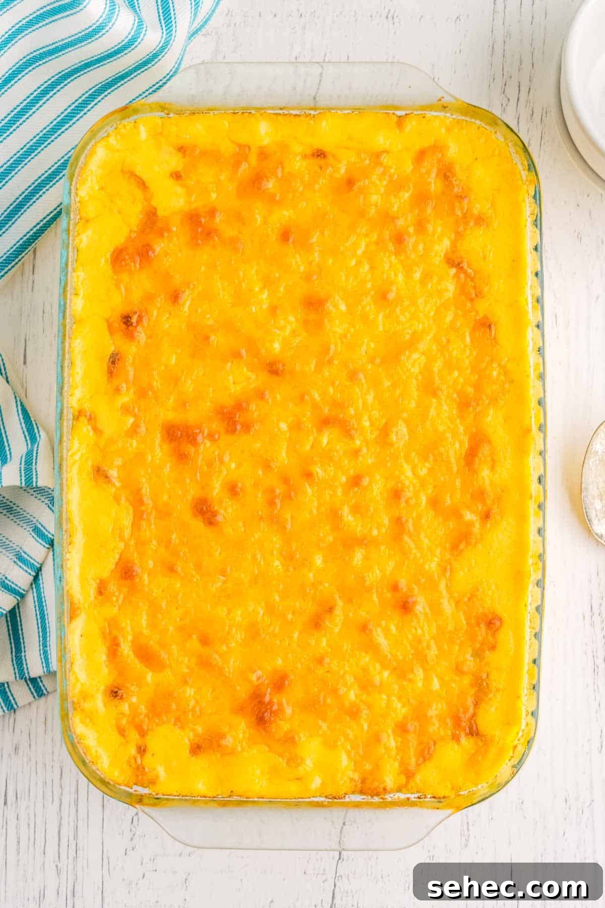 Creamy Eggless Mac and Cheese Recipe 13 A rectangular glass baking dish filled with old-fashioned mac and cheese. The surface is golden and bubbly. A striped cloth is partially visible on the left side.