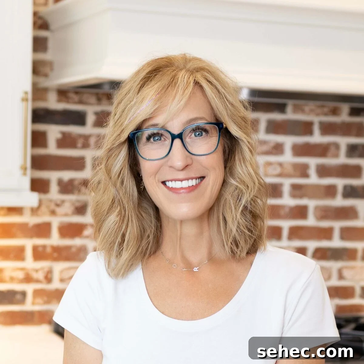 Creamy Eggless Mac and Cheese Recipe 4 Person with shoulder-length blond hair and blue glasses smiling indoors in front of a red brick wall.