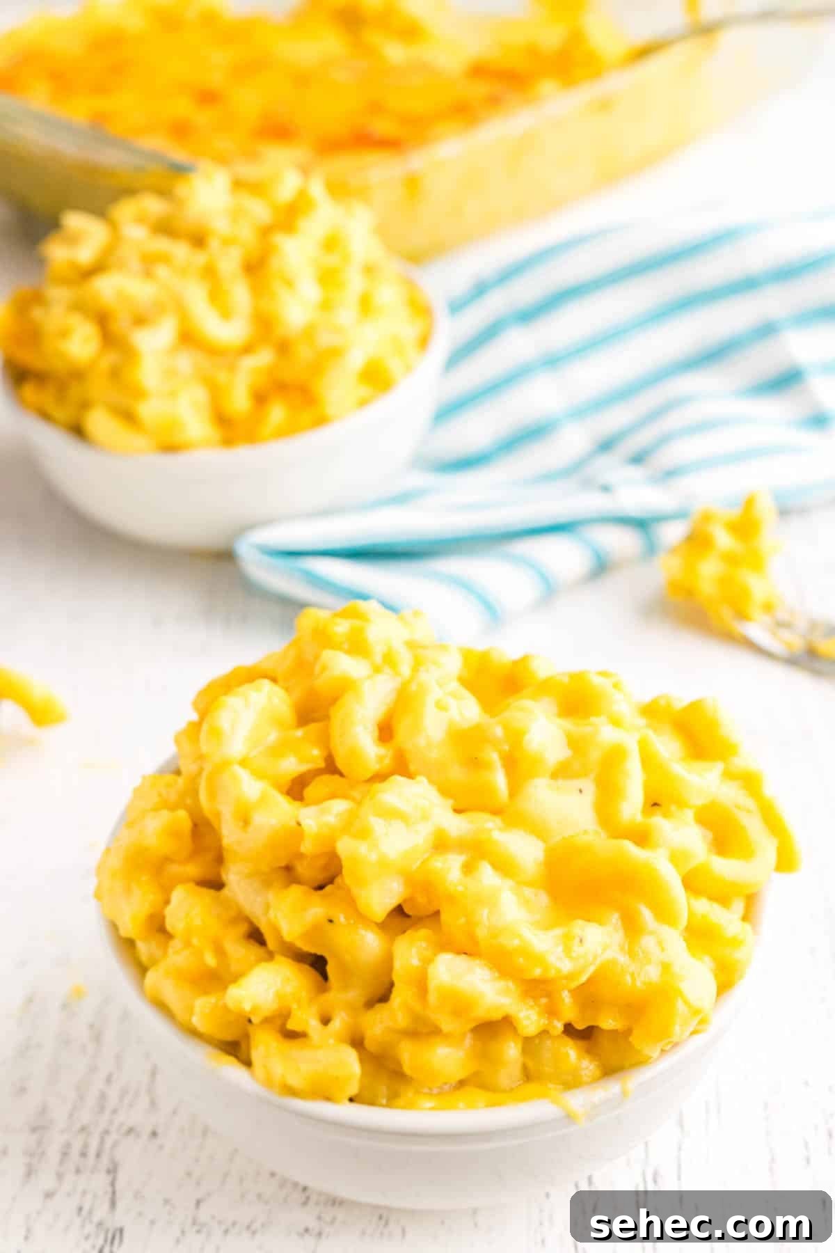 Creamy Eggless Mac and Cheese Recipe 6 Close-up of two bowls of creamy macaroni and cheese on a white surface, with a blue striped cloth and a baking dish of mac and cheese in the background.
