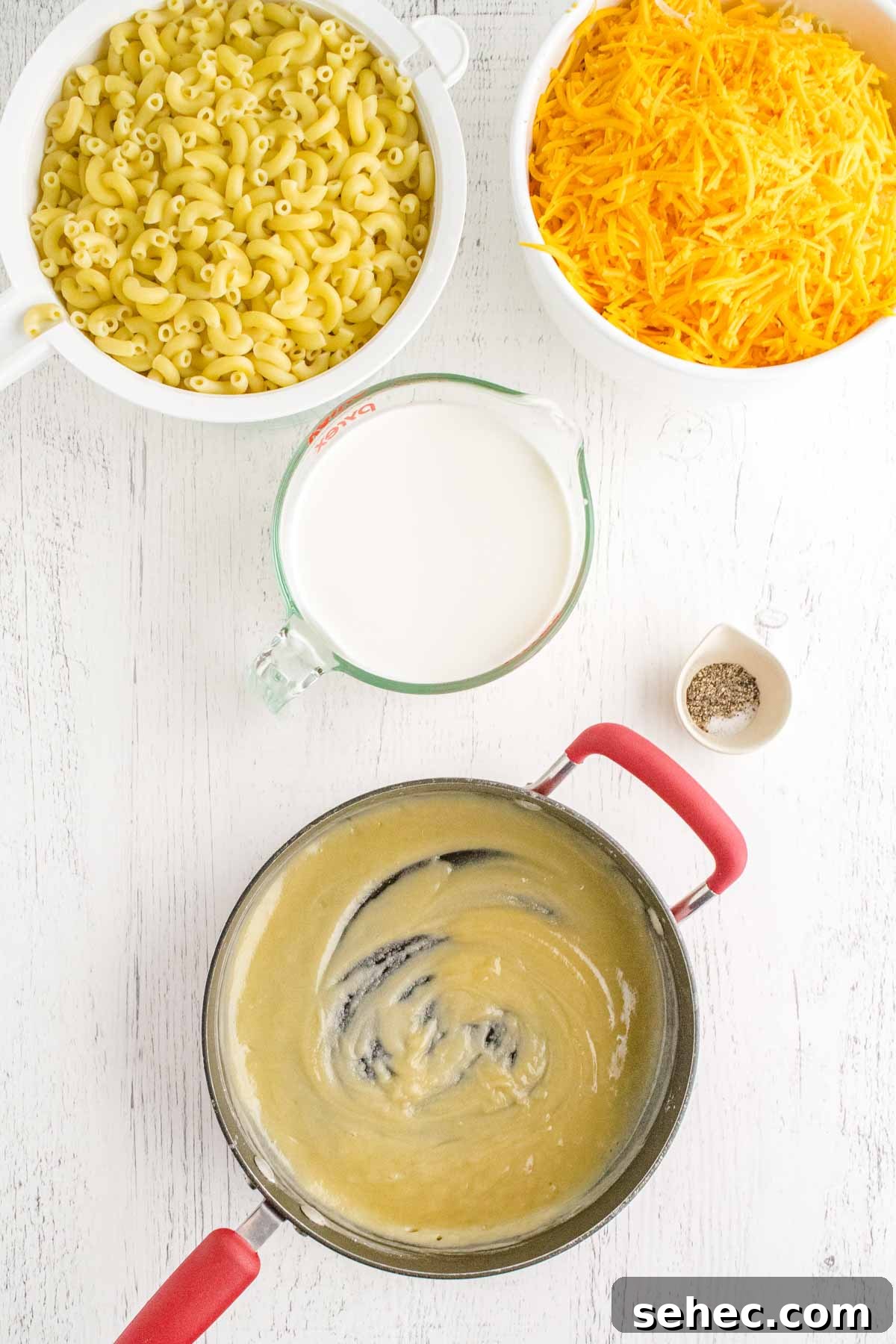 Creamy Eggless Mac and Cheese Recipe 7 Ingredients for macaroni and cheese: cooked elbow macaroni, shredded cheese, milk in a measuring cup, and a saucepan with a roux, all on a white surface with a small bowl of pepper.