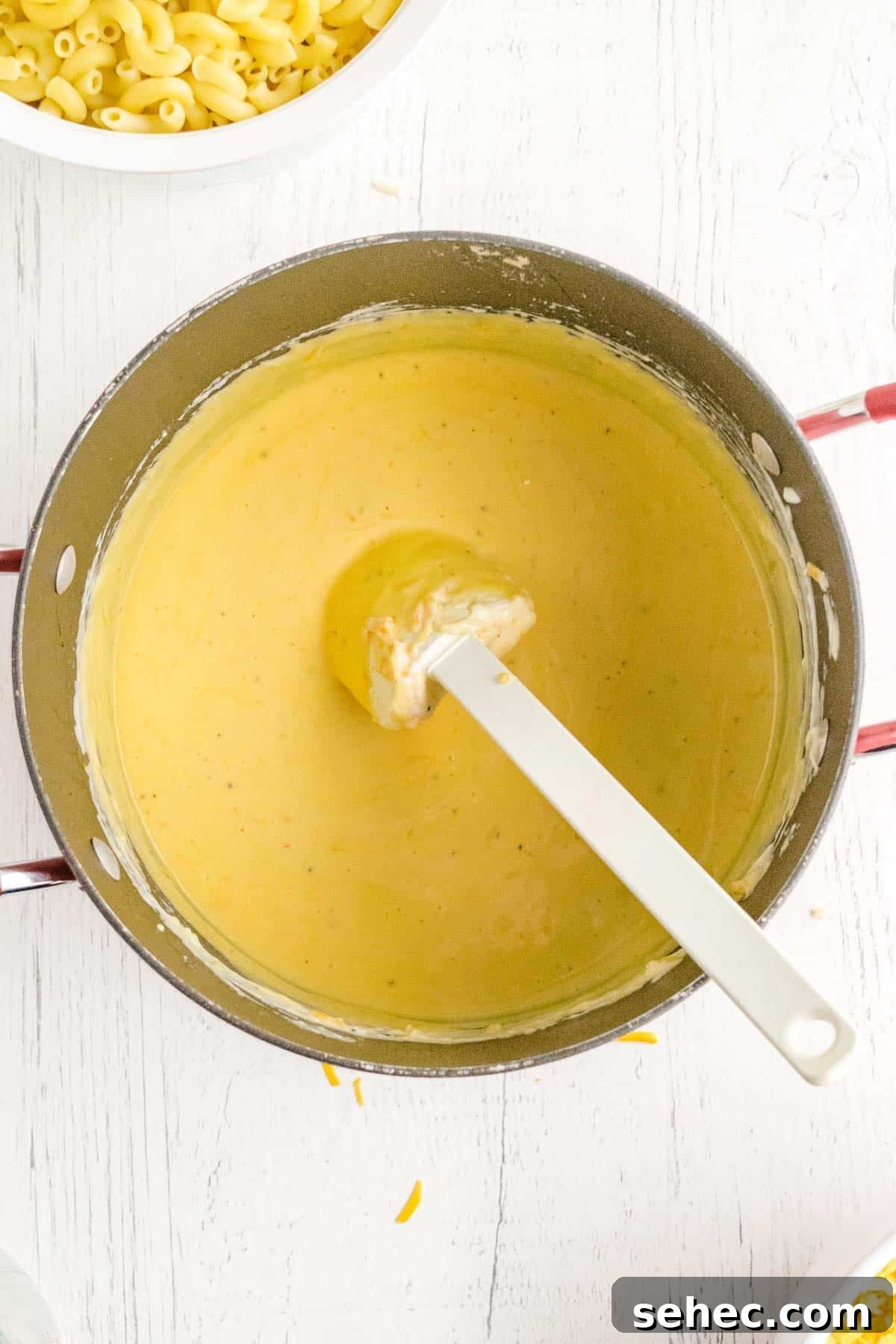 Creamy Eggless Mac and Cheese Recipe 10 A pot filled with creamy yellow cheese sauce sits on a white surface. A spatula rests in the sauce, and a bowl of uncooked elbow macaroni is visible nearby.