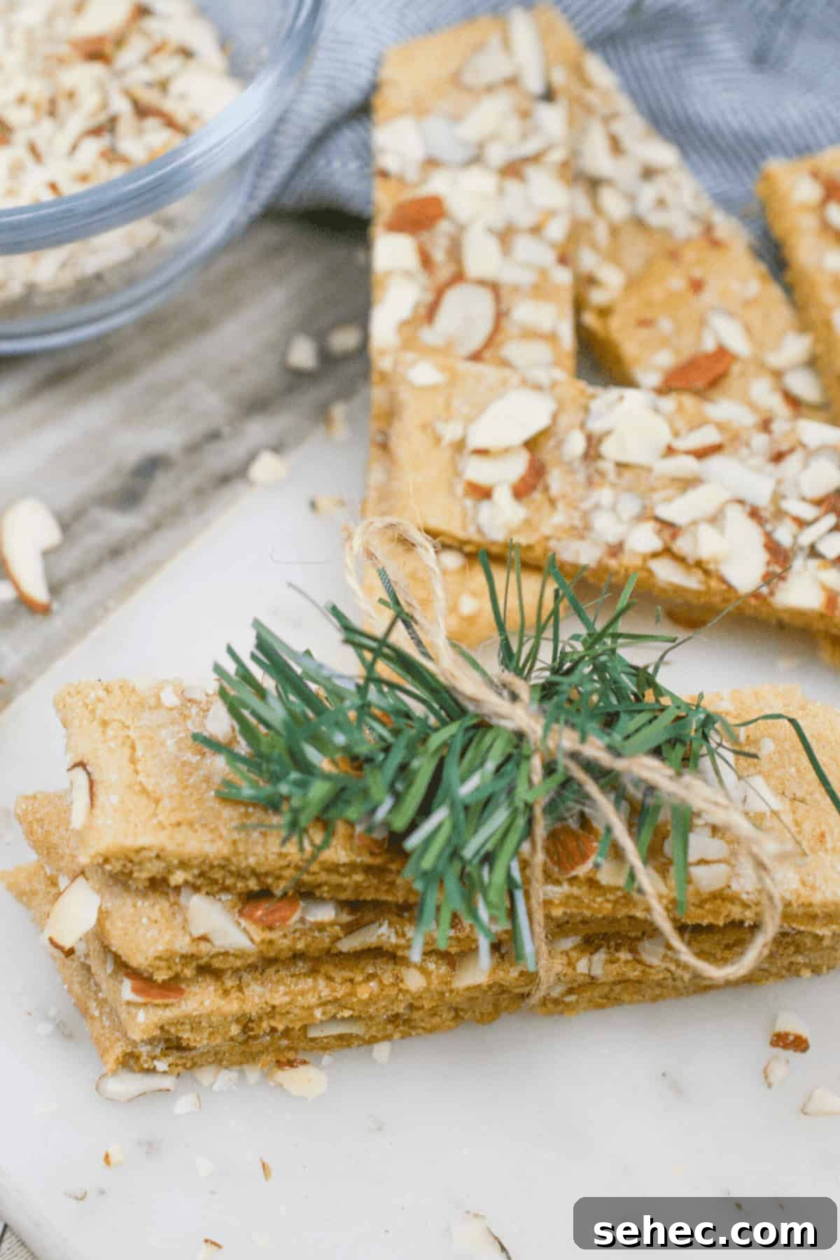 Norwegian Brune Pinner Cookies 8 Sliced desserts reminiscent of traditional Norwegian cookies, tied with twine and garnished with a pine sprig, rest elegantly on a marble surface. In the background, a bowl of sliced almonds completes the serene composition.