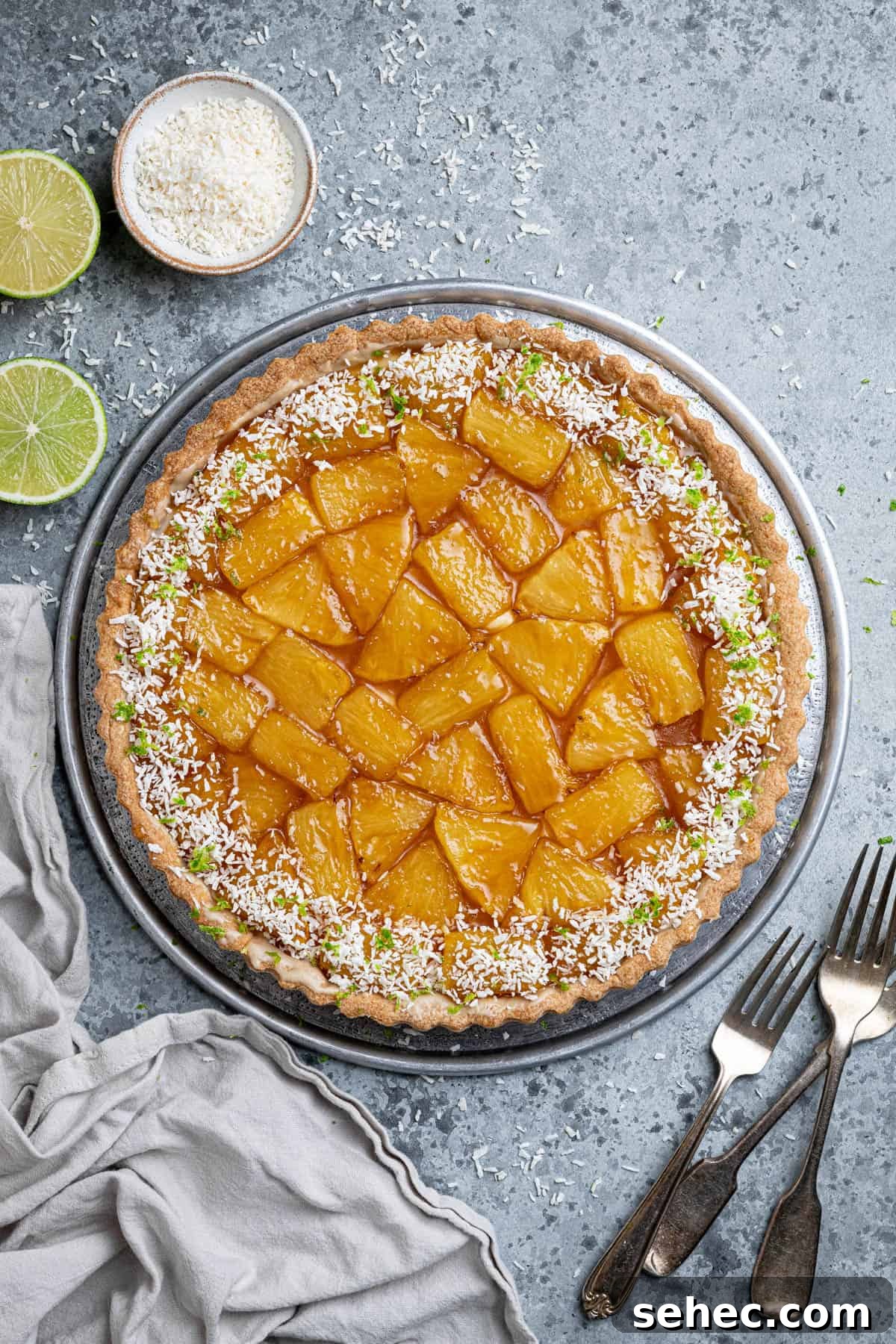 Vegan Caramelized Pineapple Coconut Tart 2 Caramelised pineapple coconut tart on a metal plate with forks, sliced limes and a bowl of desiccated coconut.