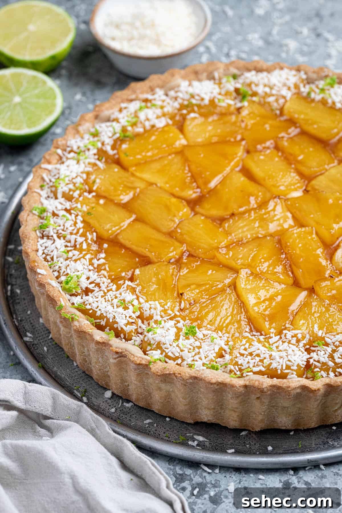 Vegan Caramelized Pineapple Coconut Tart 3 Close-up of caramelised pineapple coconut tart with sliced limes and a bowl of coconut in the background.