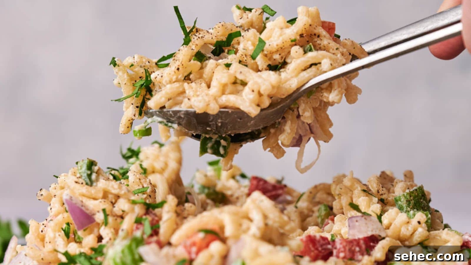 17 Budget Cookout Recipes That Save Room for Better Burgers 16 A spoon lifting a serving of creamy macaroni salad with diced red onion and fresh herbs, with pepper sprinkled on top.