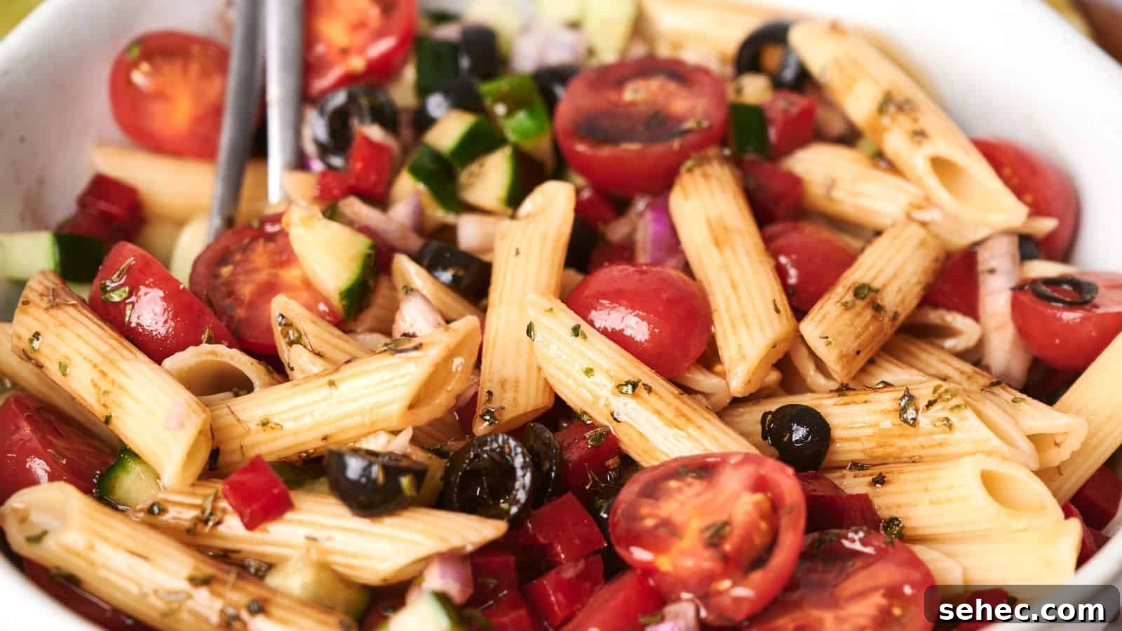 17 Budget Cookout Recipes That Save Room for Better Burgers 3 A bowl of vibrant pasta salad with penne, cherry tomatoes, black olives, diced cucumbers, red onions, and a drizzle of dressing.