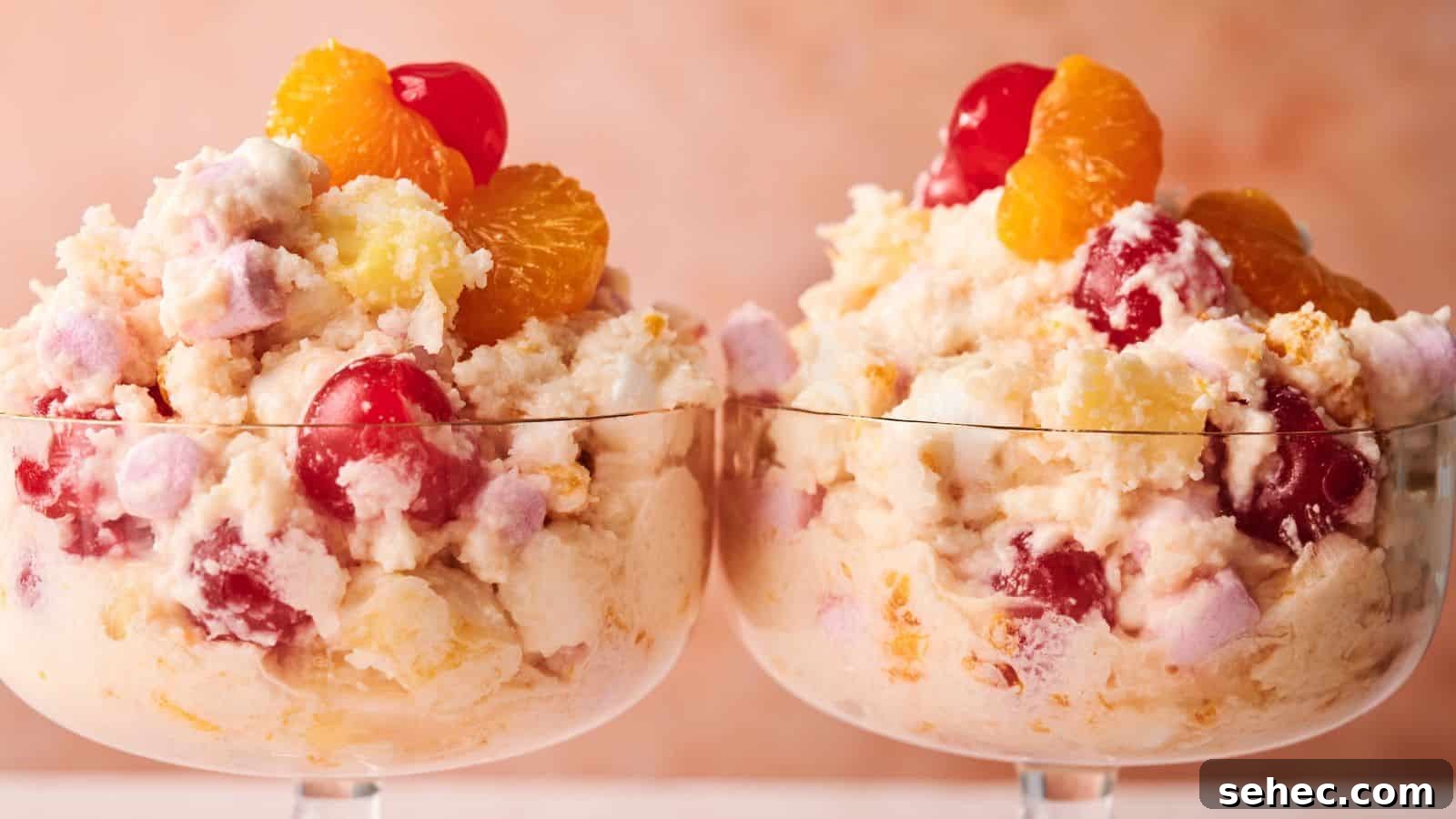 17 Budget Cookout Recipes That Save Room for Better Burgers 10 Two glass dessert cups filled with ambrosia salad, featuring whipped cream, cherries, pineapple, mandarin oranges, and marshmallows against a soft pink background.