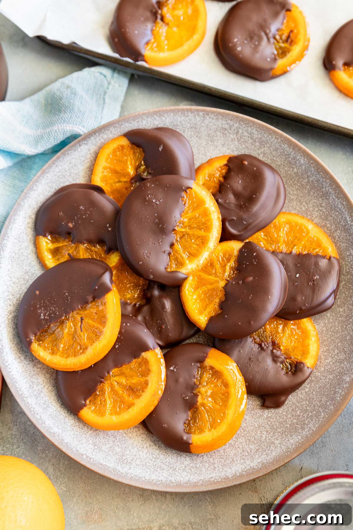 Chocolate Dipped Candied Orange Slices with Gift Tags 2 A mottled blue platter of candied orange slices dipped in chocolate with oranges and kitchen equipment around them.