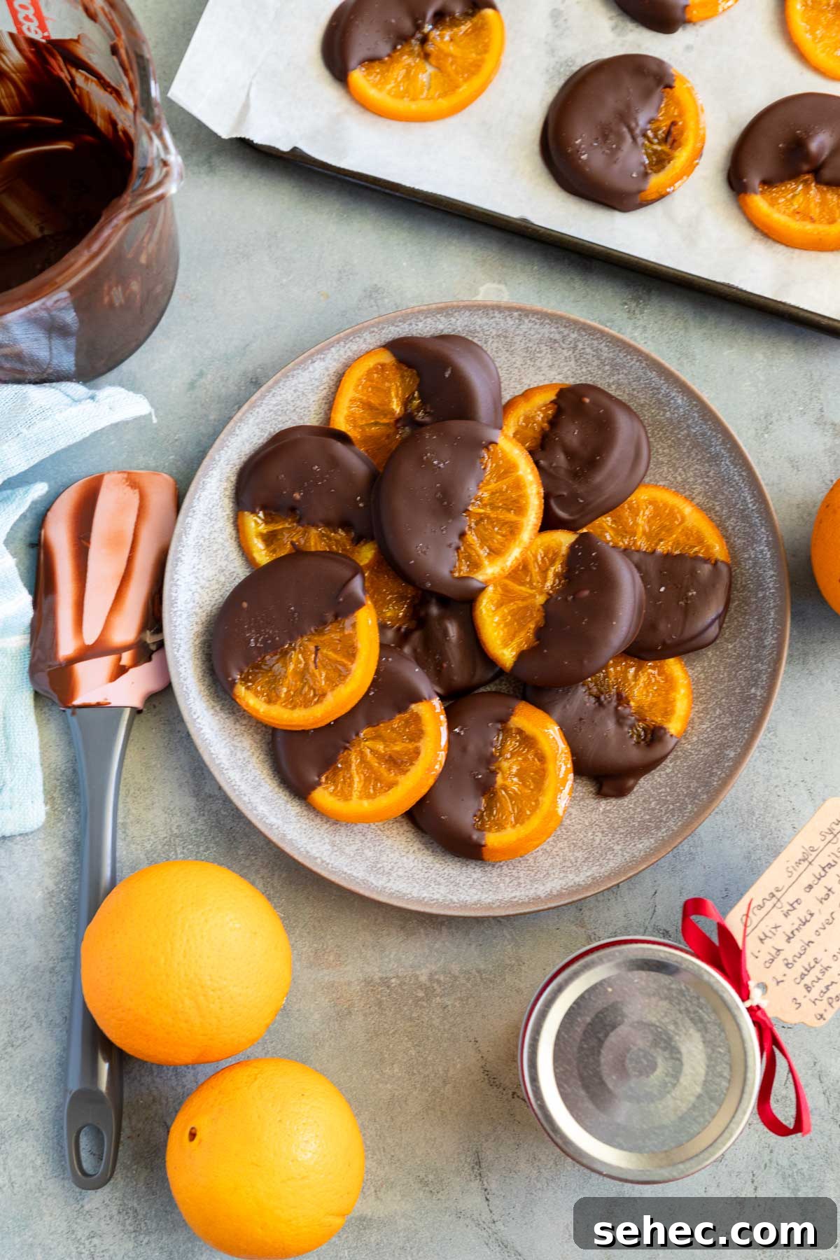 Chocolate Dipped Candied Orange Slices with Gift Tags 8 A mottled blue platter of candied orange slices dipped in chocolate with oranges and kitchen equipment around them.