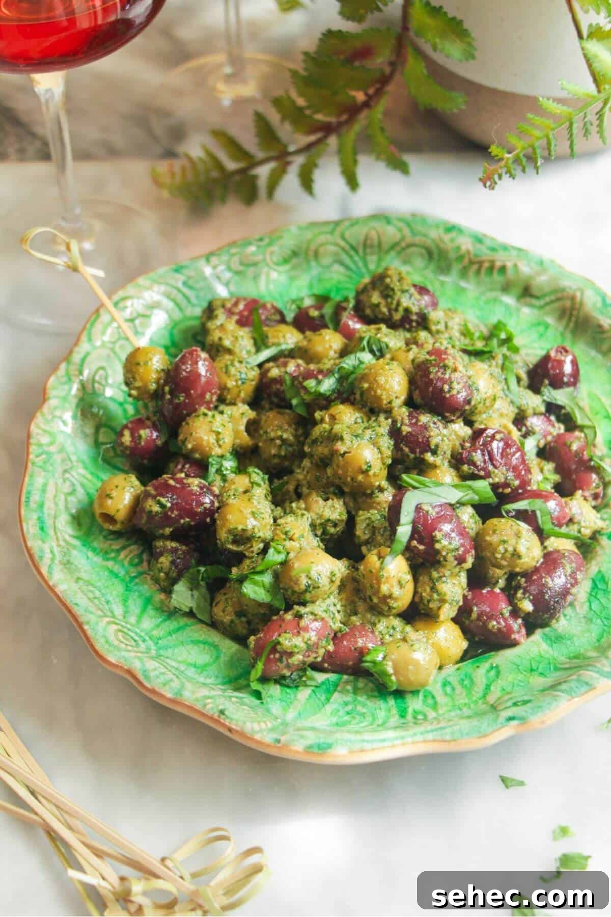 Pesto Marinated Olives Recipe 2 Pesto marinated olives on a small green plate with skewers on the side.