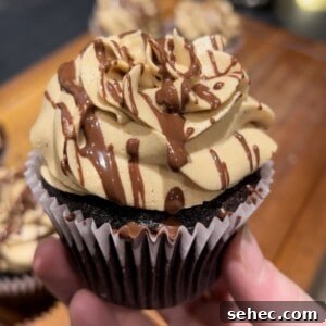 Espresso Chocolate Cupcakes with Peanut Butter Frosting 4 A hand holds a chocolate cupcake topped with swirls of light brown frosting and drizzled with chocolate. The background is blurred, showing more cupcakes on a wooden surface.