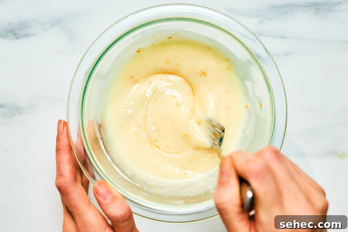 A woman's hand mixing orange flavored icing in a glass bowl.