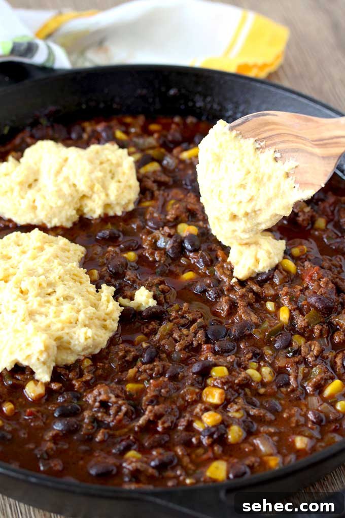 Brown Butter Cornbread Tamale Pie 4 Raw Cornbread is dolloped over a mixture of ground beef chili with beans and corn, using a wooden spoon.