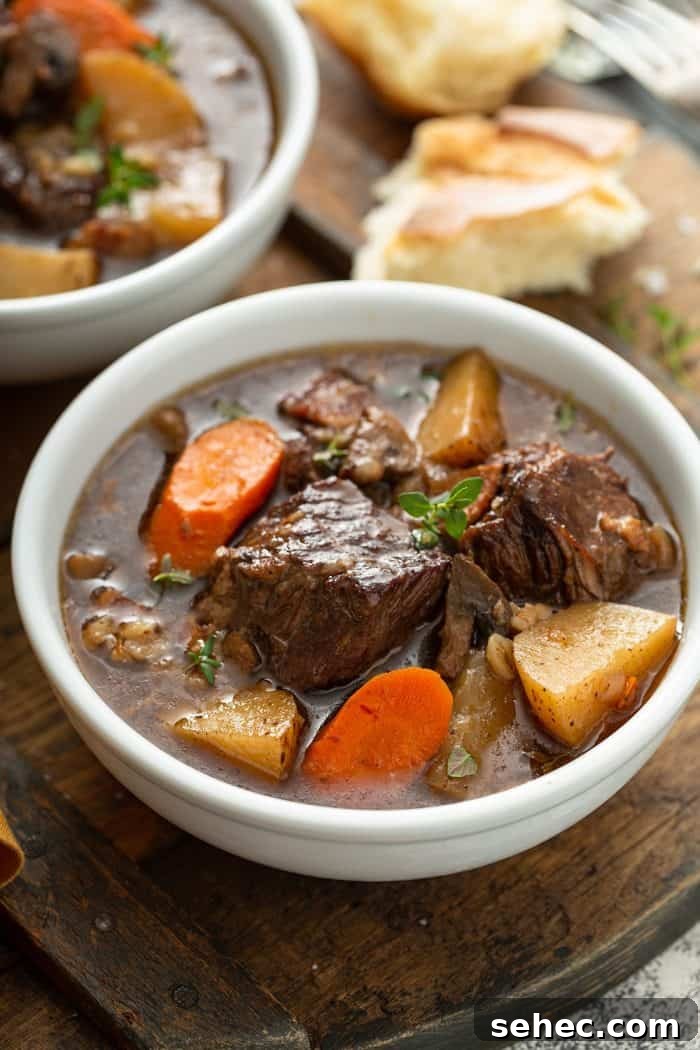Slow Cooker Beef and Barley Stew Recipe 16 Close up of a beef and barley stew in a white bowl with crusty bread in the background