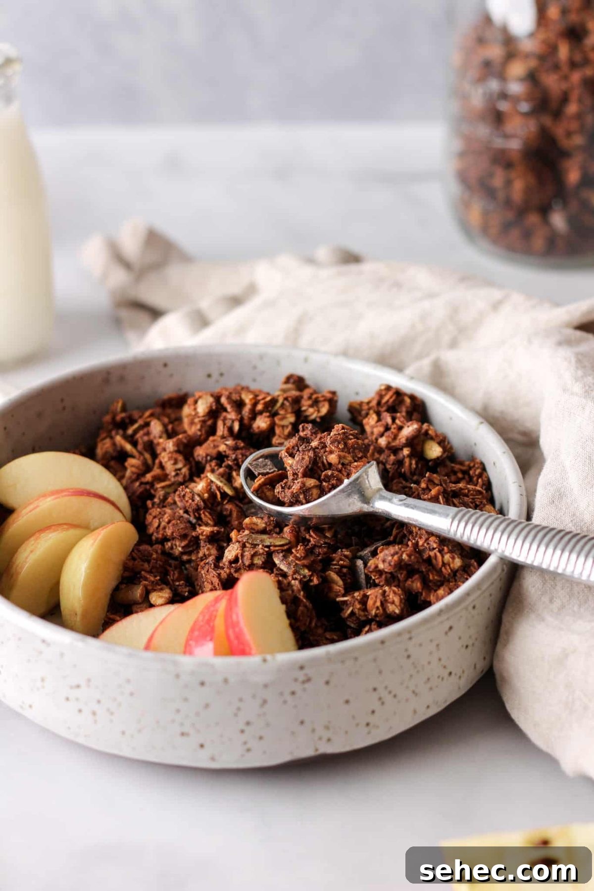 Dark Chocolate Granola Recipe a bowl of chocolate chunk granola with a spoon and sliced apples and a jar of milk