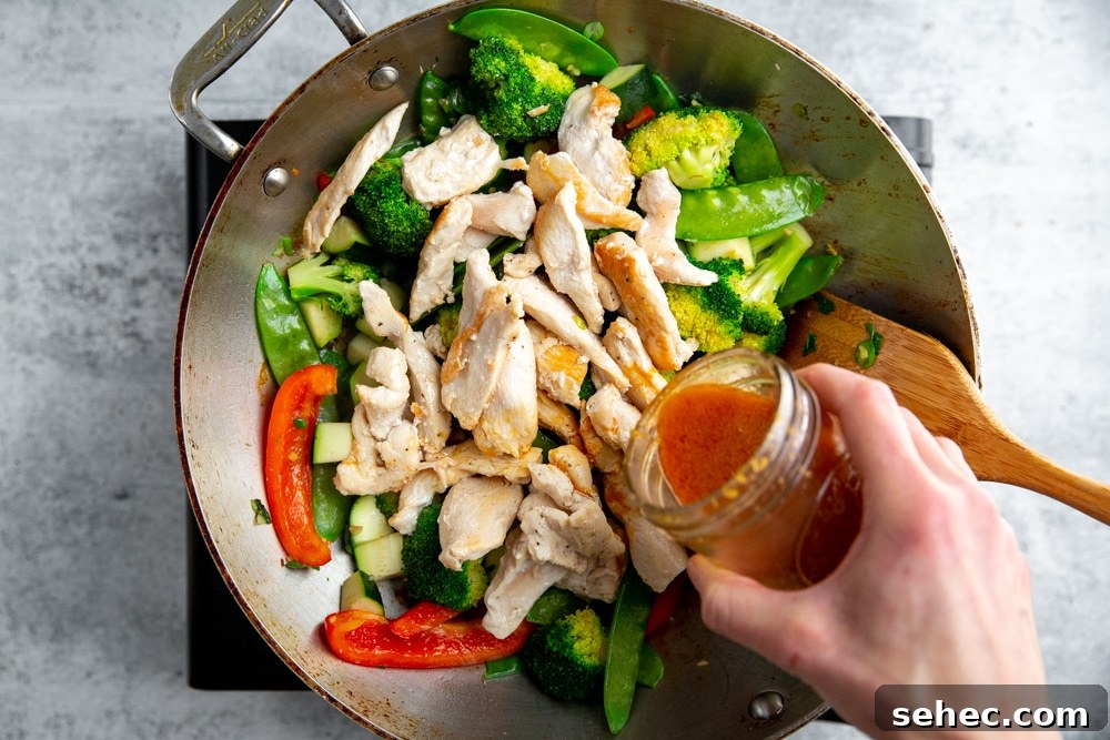Stir Fry Recipe with Any Vegetables and Meat 9 A hand pouring stir fry sauce into sauteed vegetables and chicken in a wok.