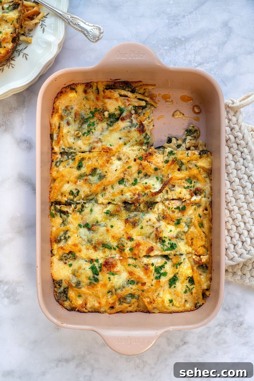 Marry Me Chicken Pasta Bake with Creamy Tuscan Sauce 2 A Marry Me Chicken Pasta Bake casserole topped with melted cheese and garnished with fresh herbs sits in a rectangular dish on a marble countertop. A portion has been served on a nearby plate, revealing the dish's textured and creamy interior.