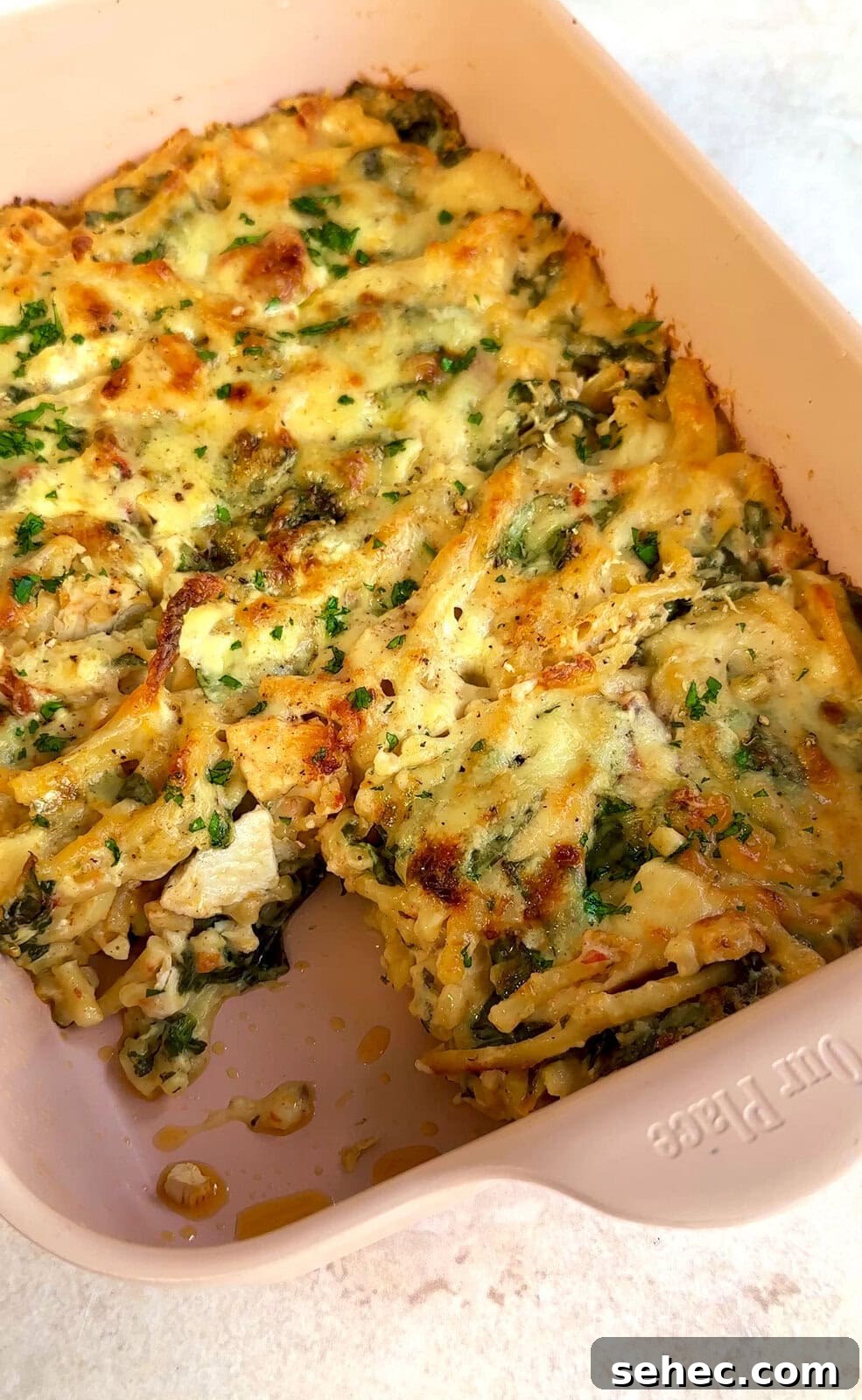 Marry Me Chicken Pasta Bake with Creamy Tuscan Sauce 3 A baked casserole in a rectangular dish, featuring golden brown, melted cheese and mixed herbs on top. The dish is partially cut, revealing layers of pasta and visible greens.
