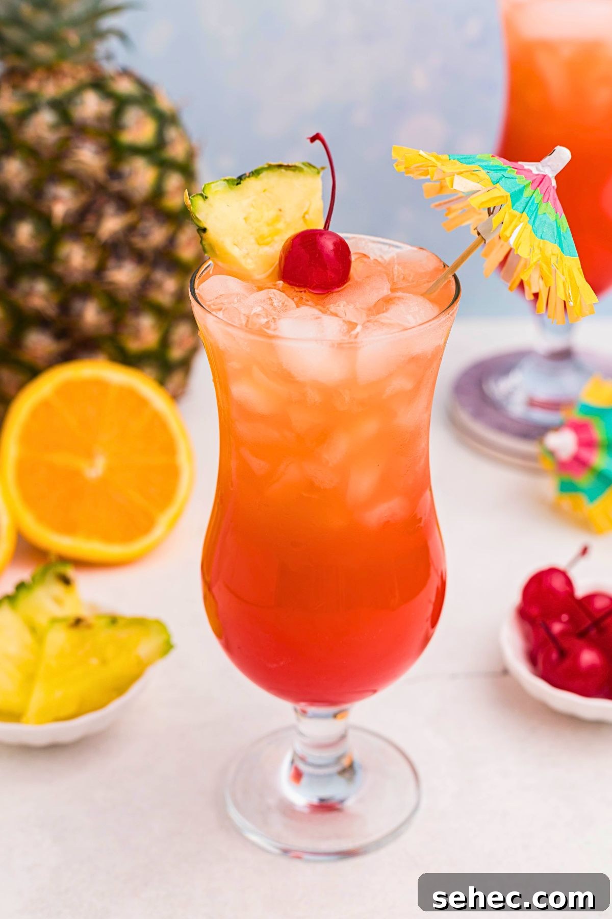 Bahama Mama Cocktail Recipe 2 Red and orange Bahama Mama cocktail in a hurricane glass with orange slices, cherries, and a paper umbrella on a marble table.