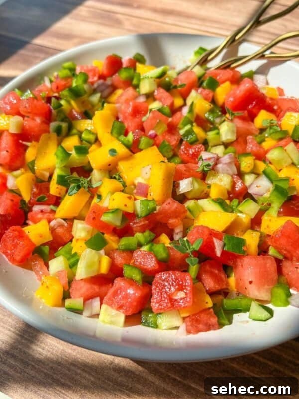 A close-up of a colorful salad with diced watermelon, mango, cucumber, green bell pepper, and red onion in a white bowl, with two gold forks resting on the edge. Sunlight highlights the vibrant ingredients.