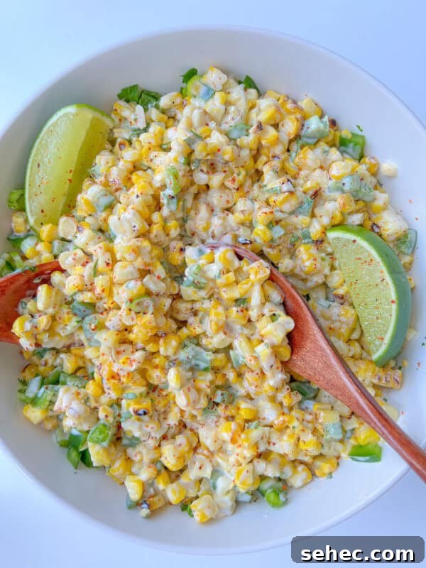 A white bowl filled with creamy Mexican Corn Salad, garnished with chopped green herbs, two lime wedges, and sprinkled with chili powder. Two wooden serving spoons are resting in the salad.