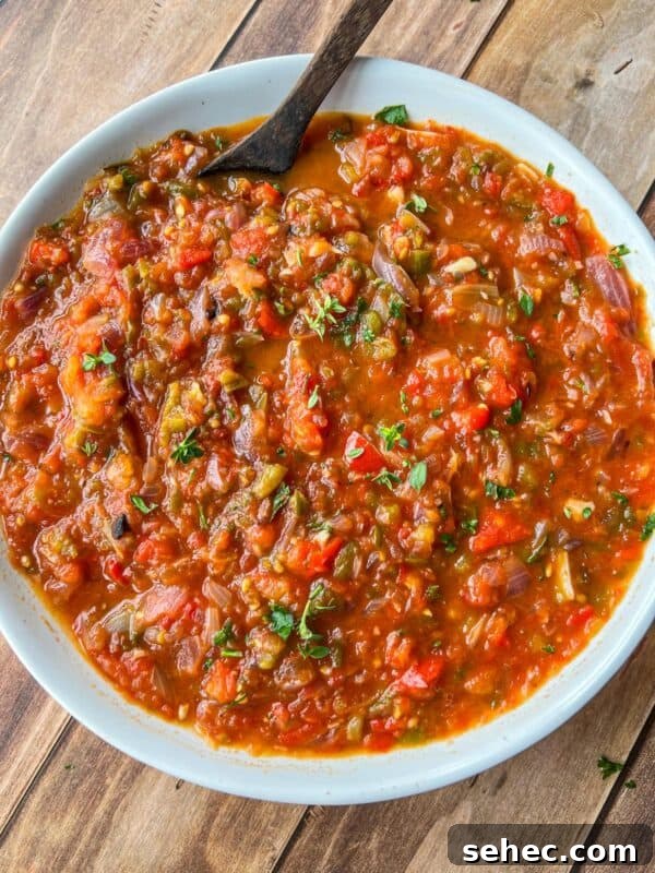 A white bowl filled with chunky, red and green salsa garnished with chopped herbs sits on a wooden surface, with a dark spoon resting in the bowl.