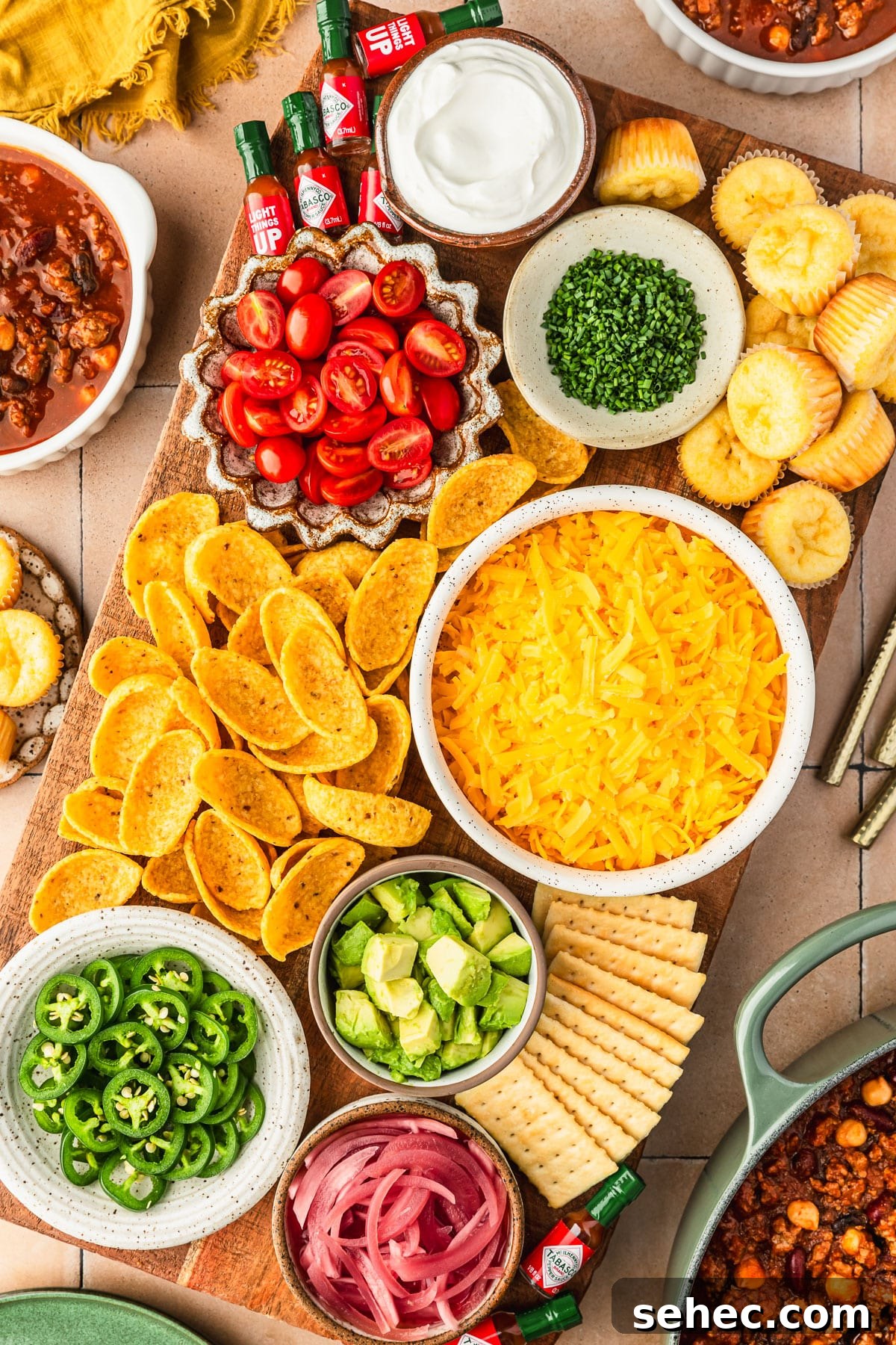 Chili Charcuterie Board with Homemade Chili Recipe 2 A chili toppings charcuterie board with white and brown bowls of cheese, sour cream, veggies, chips, and hot sauce bottles on a beige tiled counter next to bowls of chili, a green pot of chili, and a yellow linen.