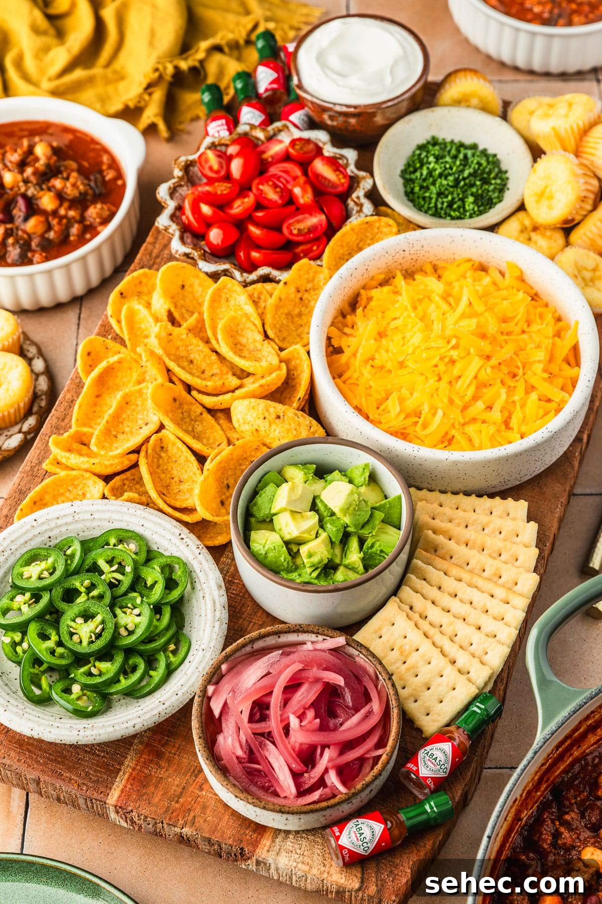 Chili Charcuterie Board with Homemade Chili Recipe 10 A chili topping charcuterie board with bowls of cheese, veggies, chips, crackers, and herbs on an orange tiled counter next to a pot of chili, bowls of chili, and yellow napkin.