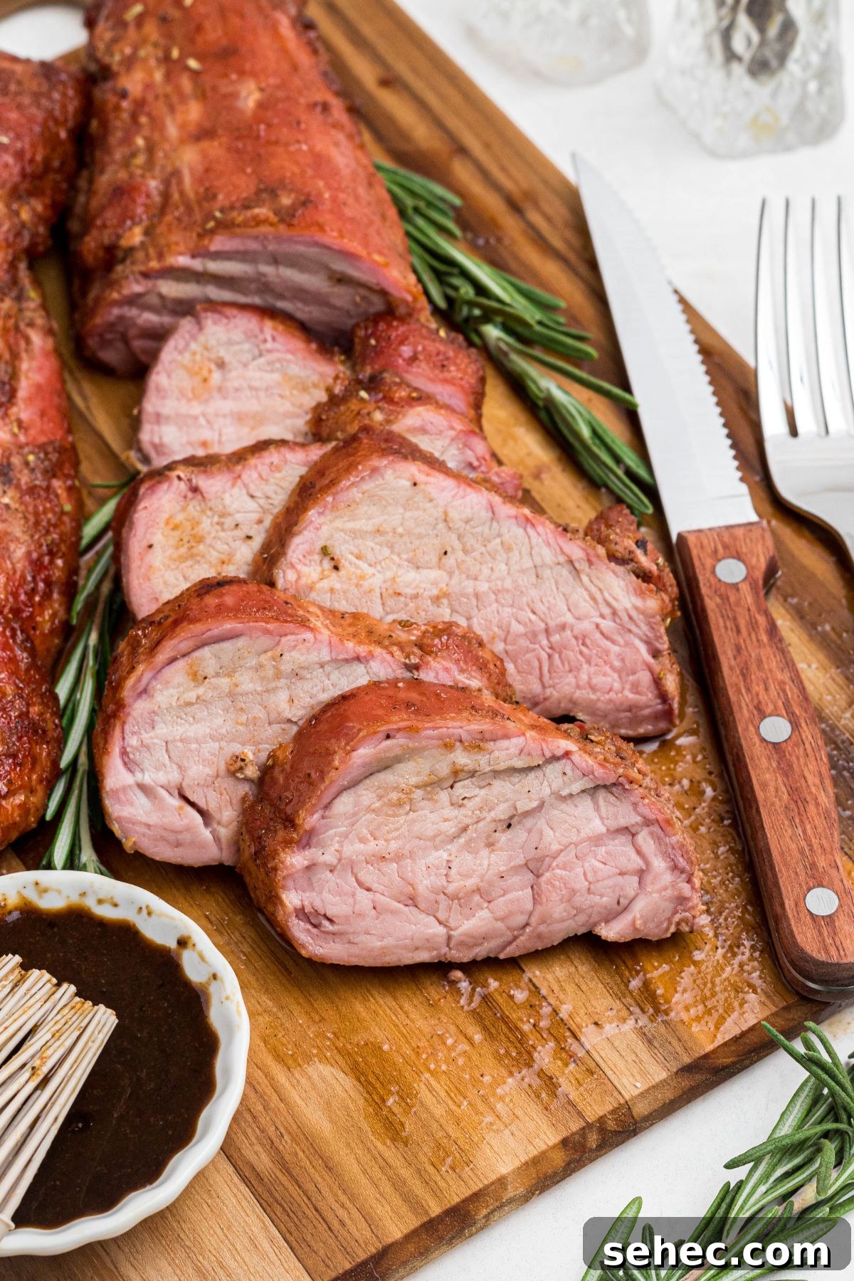 Juicy Smoked Pork Tenderloin Recipe 2 Juicy tenderloin cut into slices on a cutting board with a knife and fork, and fresh rosemary.