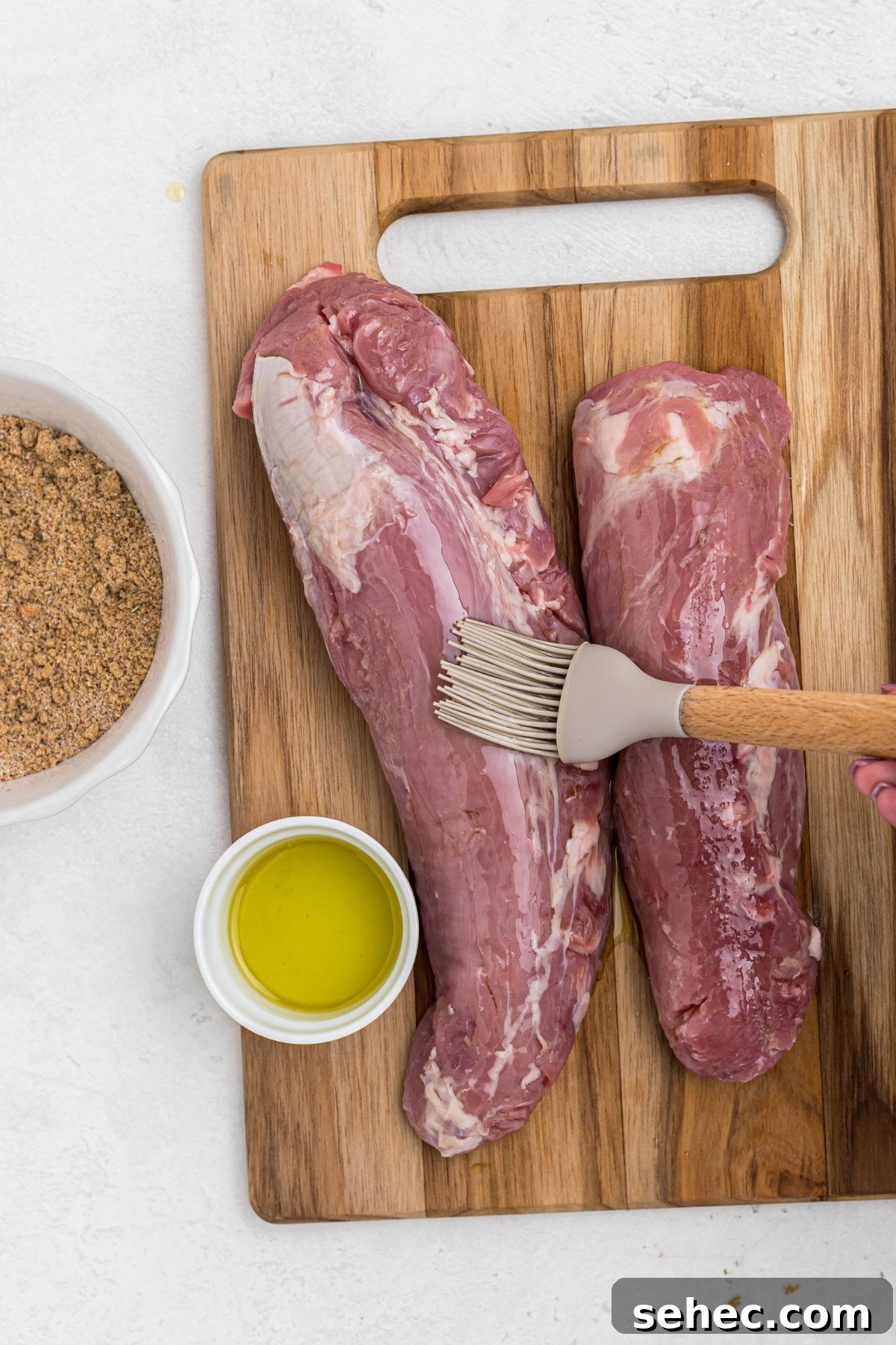 Juicy Smoked Pork Tenderloin Recipe 4 Two uncooked pork tenderloins on a wooden cutting board with olive oil being brushed on top.