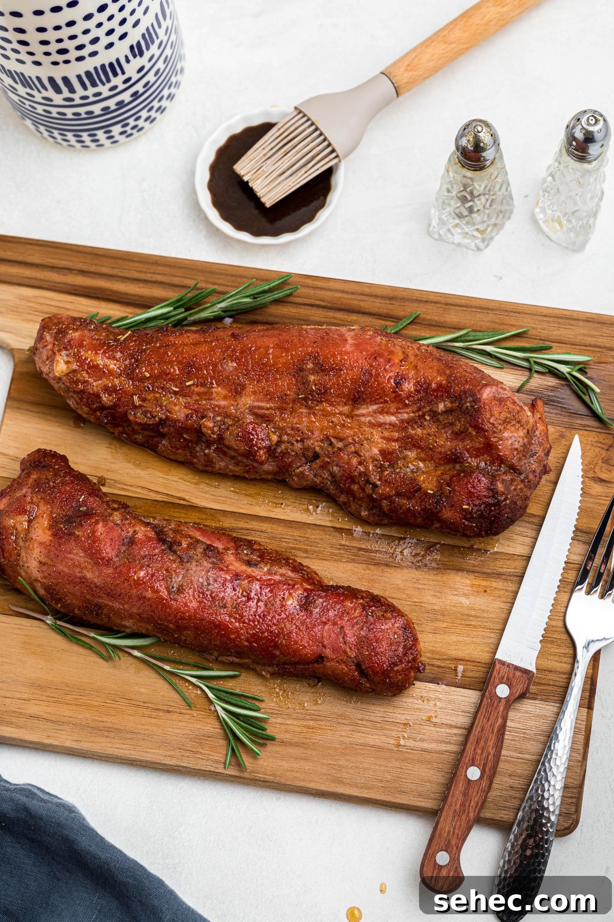 Juicy Smoked Pork Tenderloin Recipe 9 Smoked tenderloins on a wooden cutting board, resting, with sauce being brushed on the side.