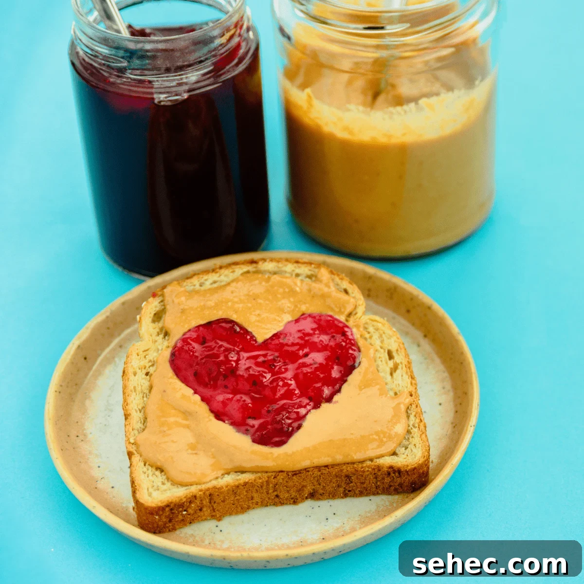 Blue background with glass jars of jelly and peanut butter behind a plate with a piece of bread covered in peanut butter with a jelly heart.